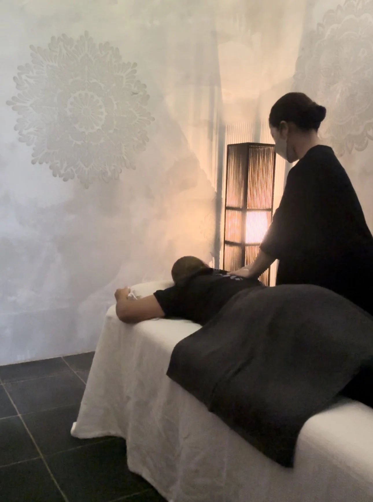 マッサージ師が女性の背中を揉んでいるマッサージベッドの上の女性と、間接照明のある暗い室内の景色