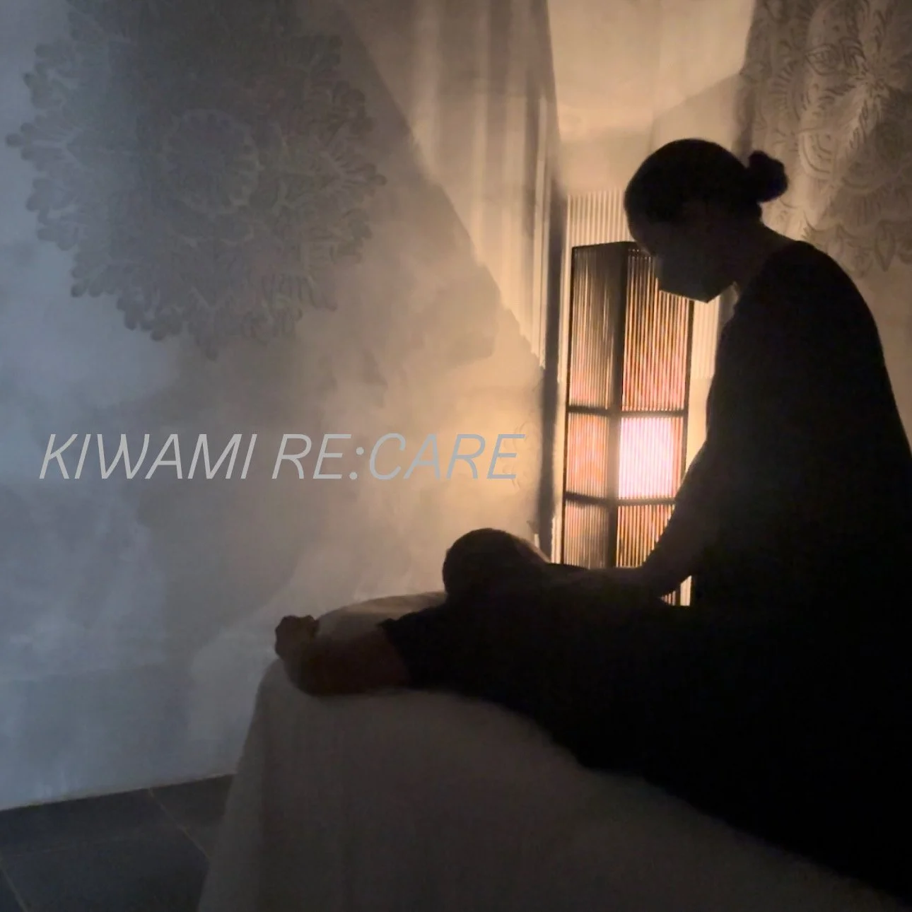 マッサージ師が顧客にマッサージを施している様子。背景には禅の草木と灯りが見える静かな室内の空間。