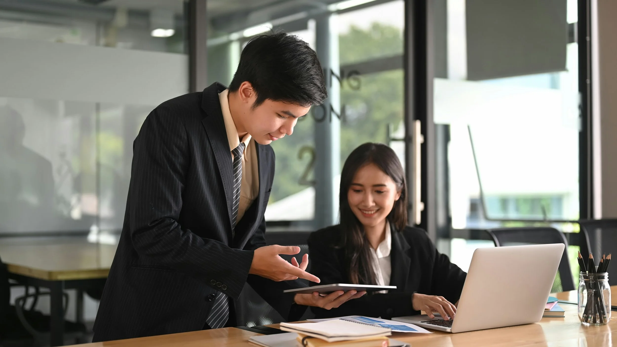 A man in a black suit showing something on a tablet to a woman in a black blazer, seated at a desk with a laptop, papers, and pencils in a modern office.