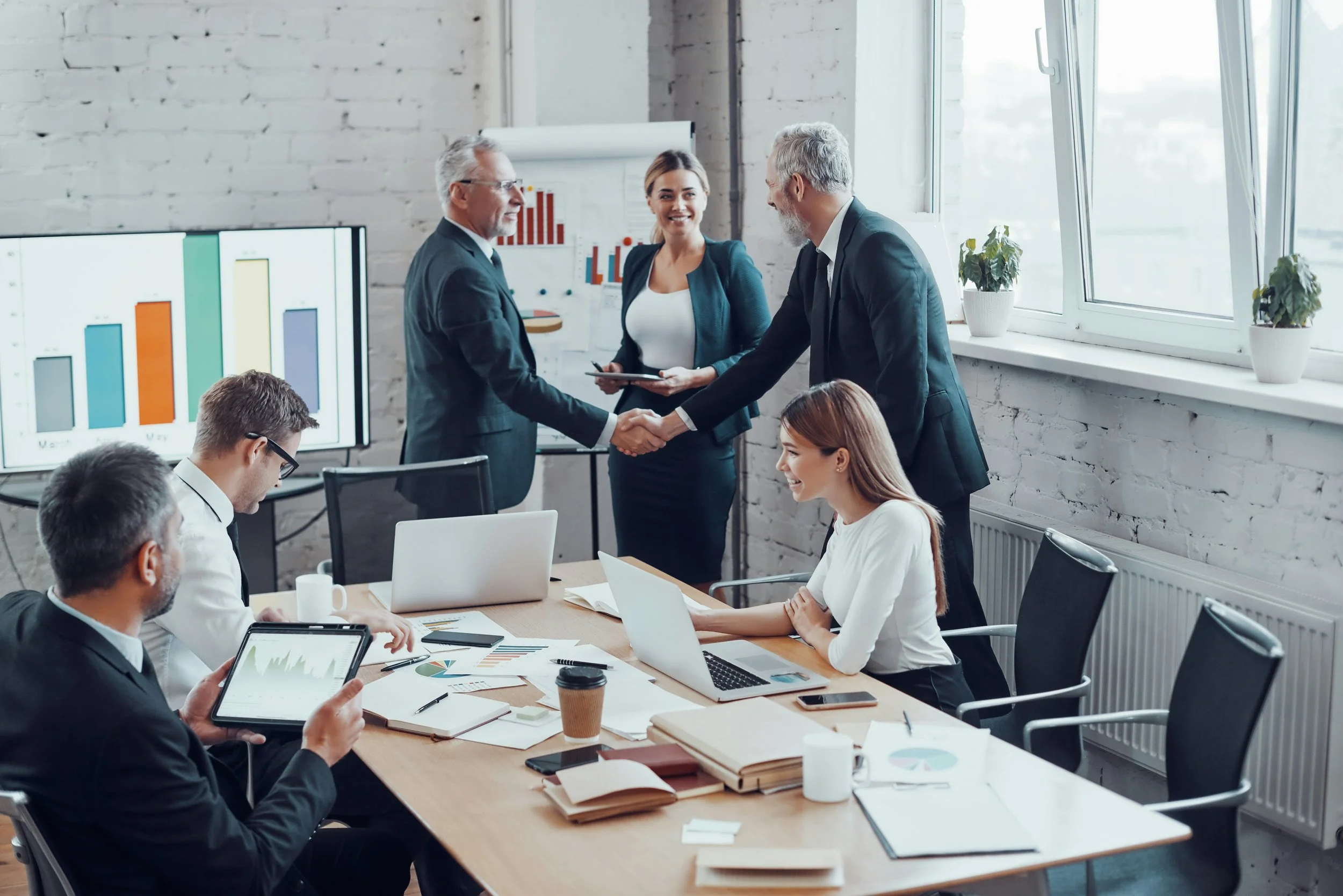 Businesspeople in a meeting room shaking hands while others observe and take notes.