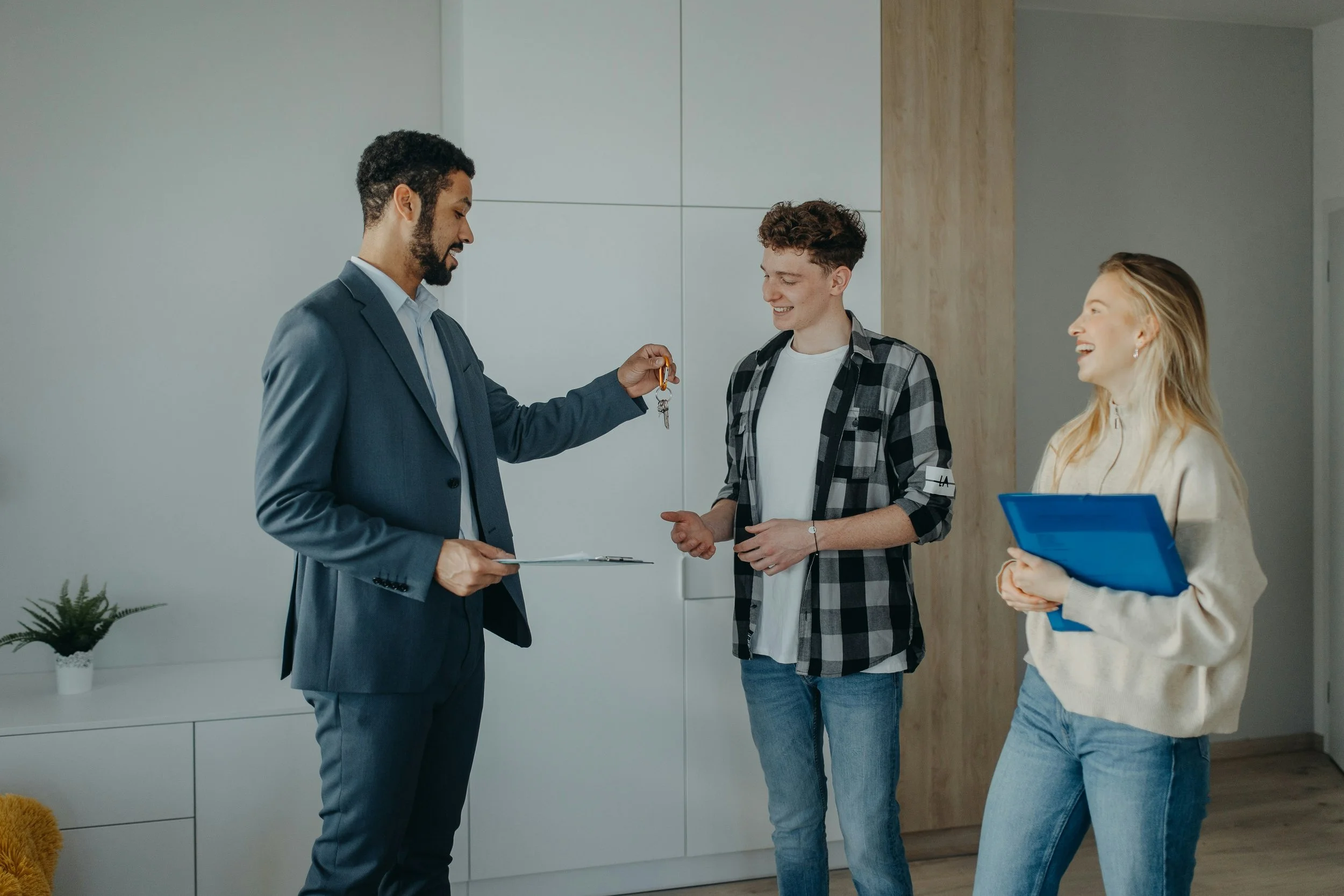 A man in a suit hands over car keys to a young man, while a young woman holds a blue folder, all smiling inside modern office or home with minimal decor.