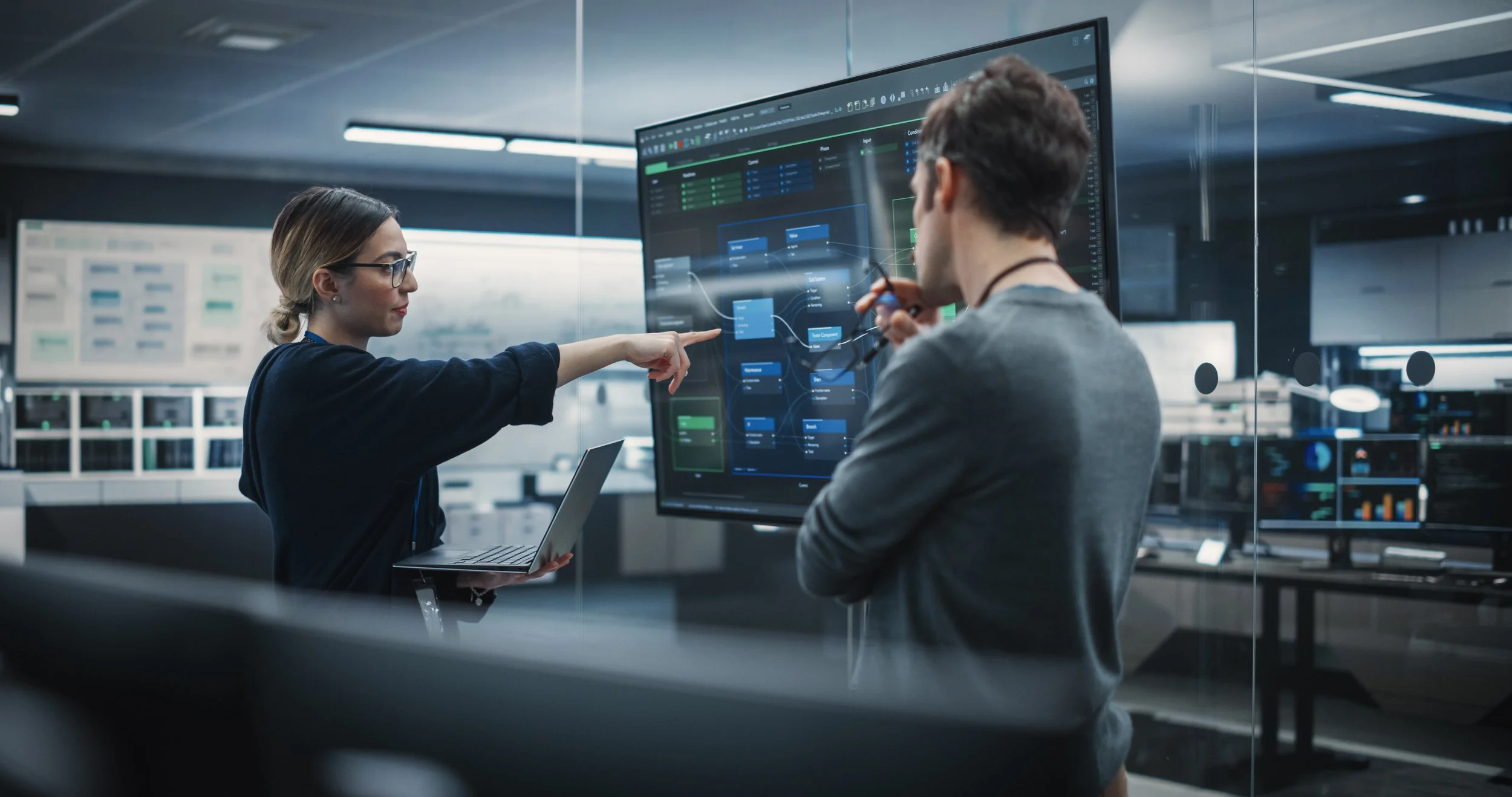 Two people in a modern office, one woman and one man, discussing data on a large digital display with flowcharts and graphs, while the woman holds a laptop.