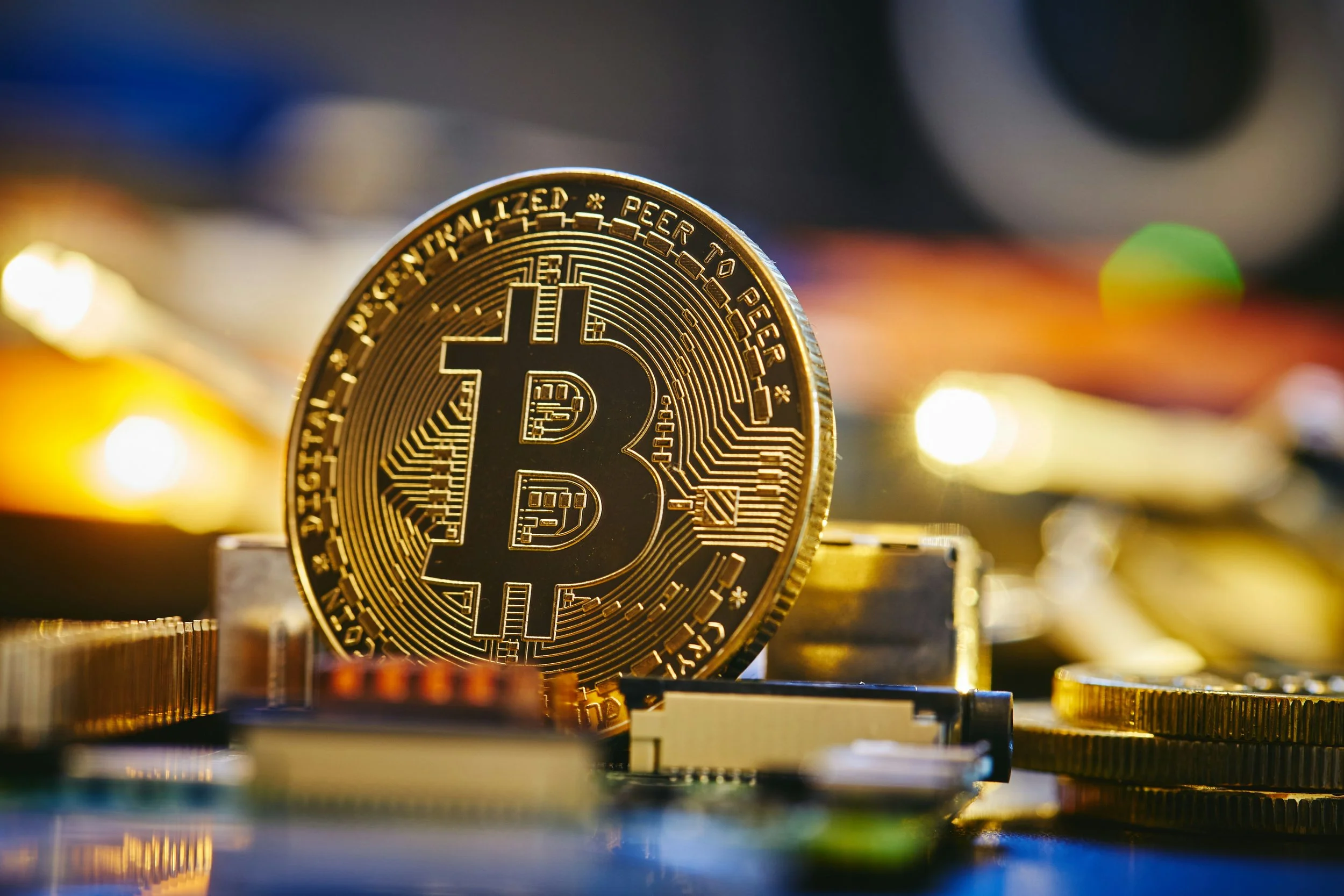 Close-up of a physical Bitcoin coin standing on a surface, with other coins and electronic components in the blurred background.