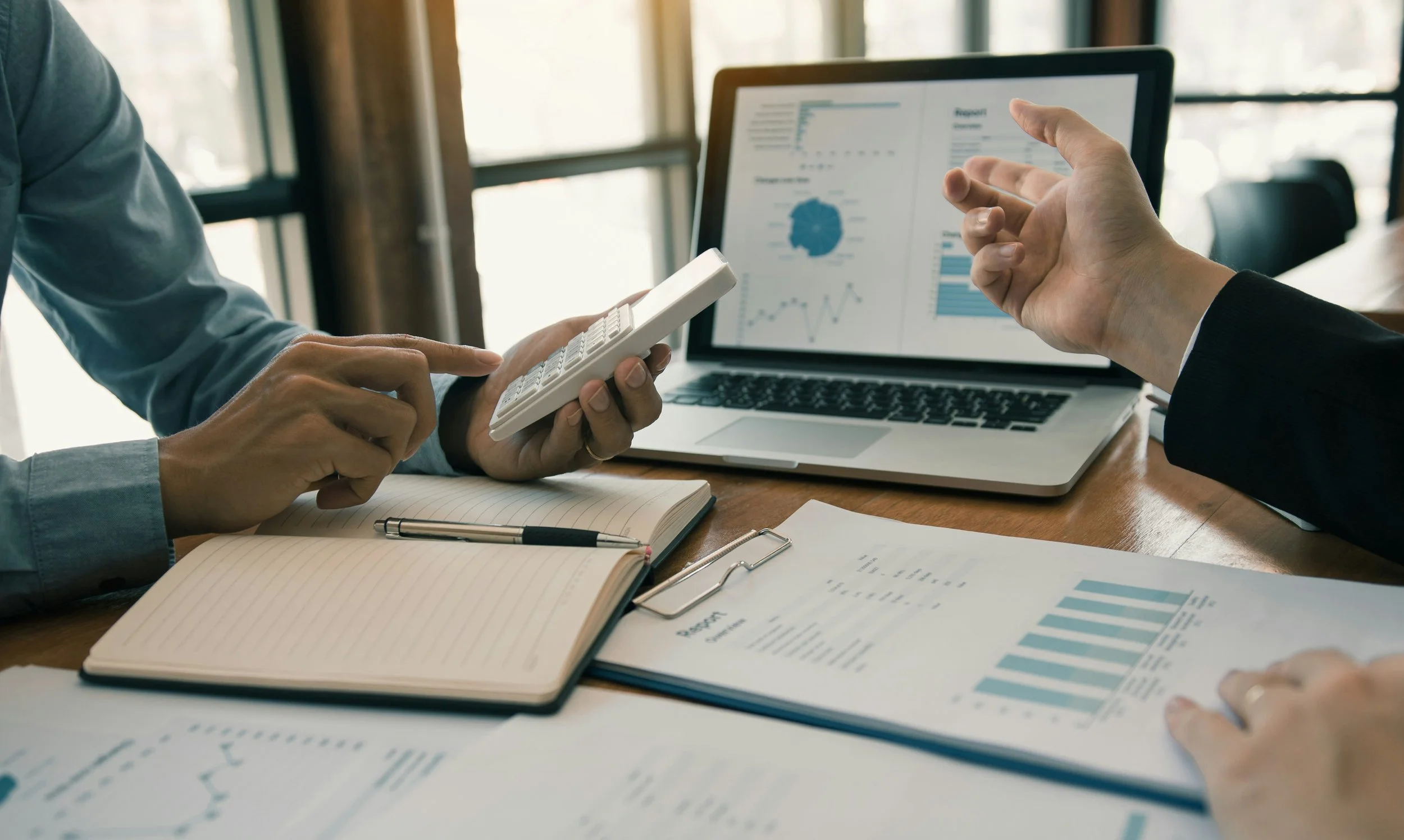Two people are discussing financial data at a desk with a laptop, notebook, and charts, in a well-lit office.
