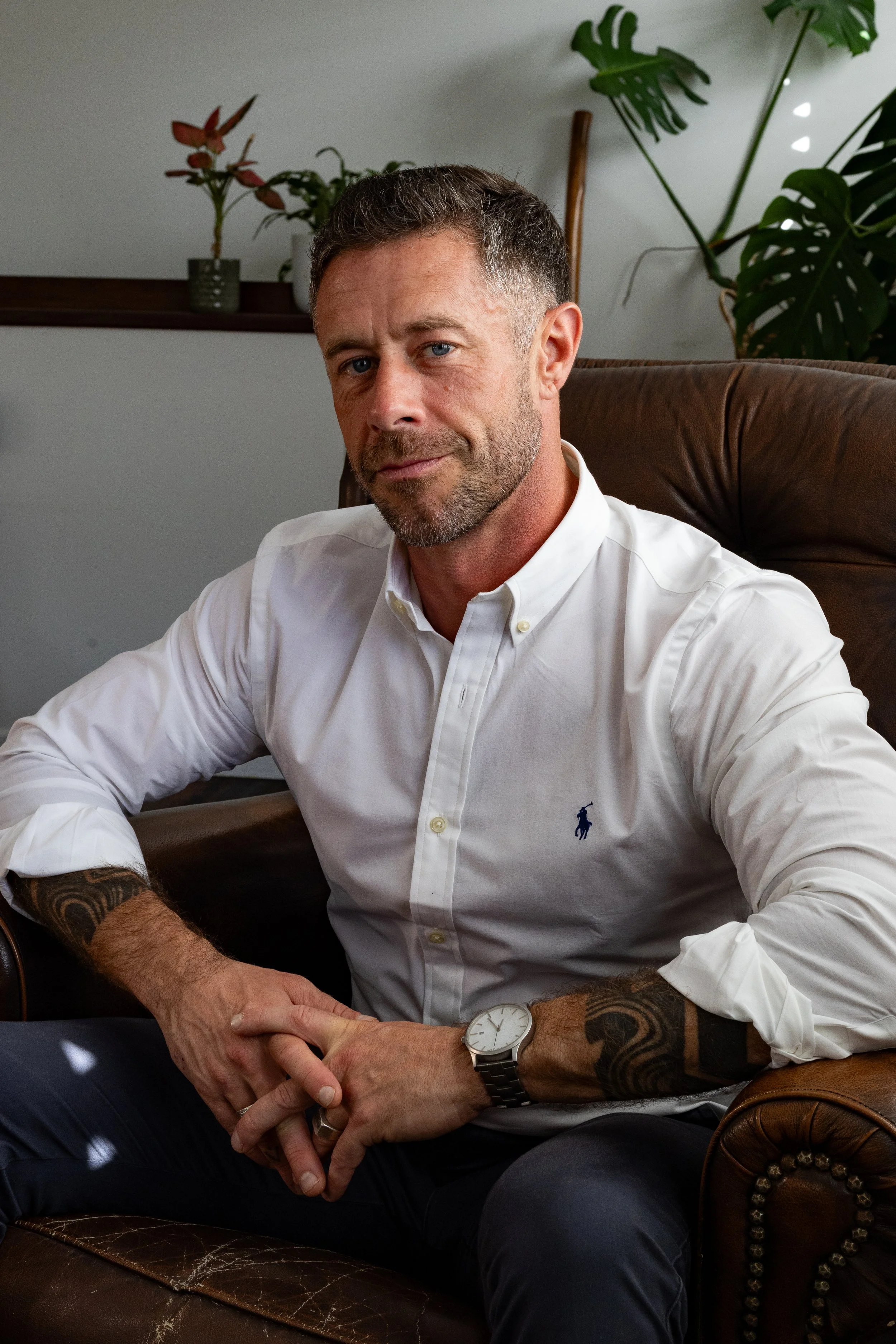 A man with short brown hair, blue eyes, and a beard sitting on a brown leather armchair, wearing a white button-up shirt, a watch, and tattoos on his left arm.
