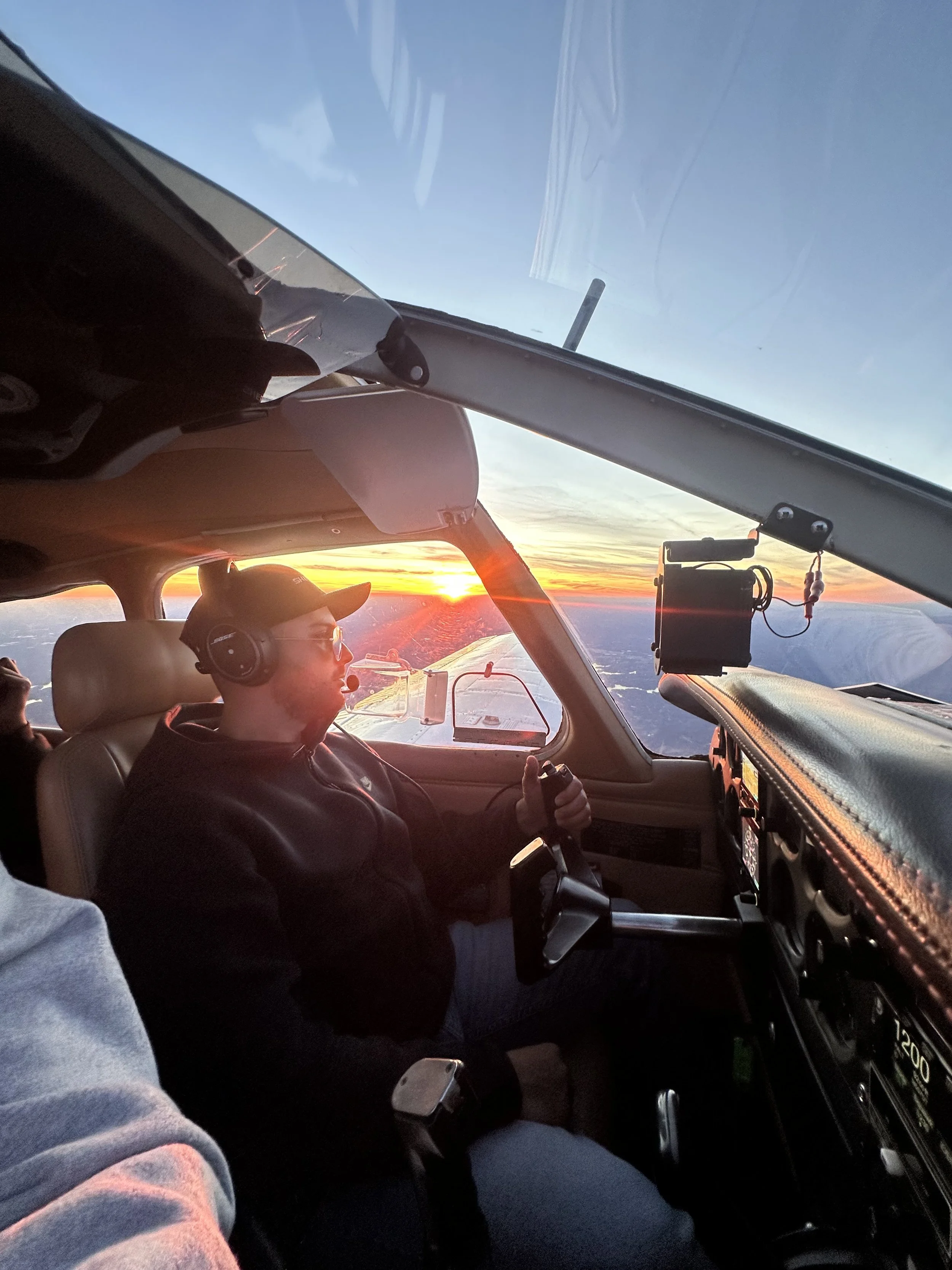 Pilot flying a small airplane during sunset, with the pilot wearing headphones and sunglasses, overlooking a landscape with a colorful sky.