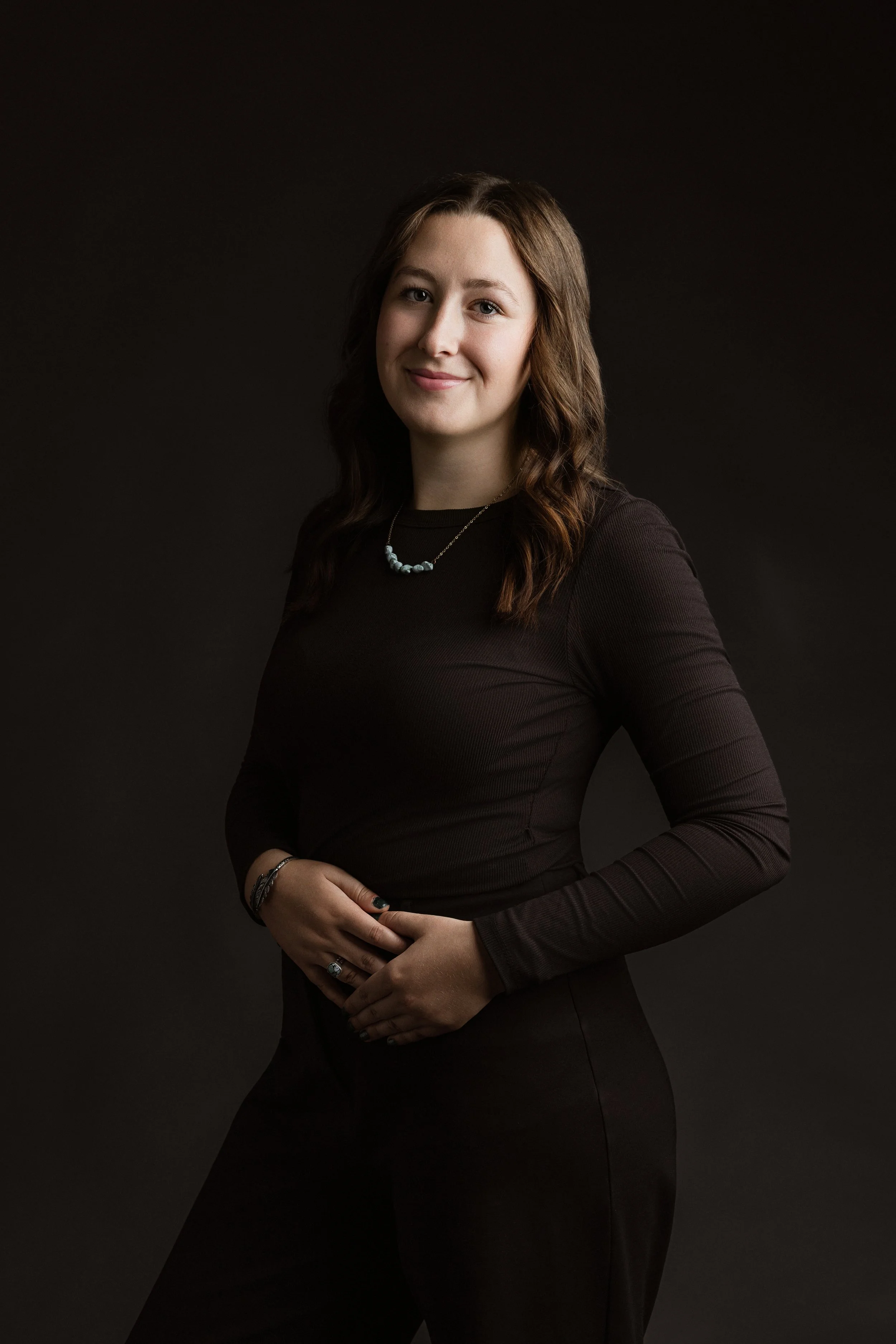 A woman with wavy brown hair wearing a dark long-sleeve top, a beaded necklace, and rings, standing against a dark background and smiling at the camera.