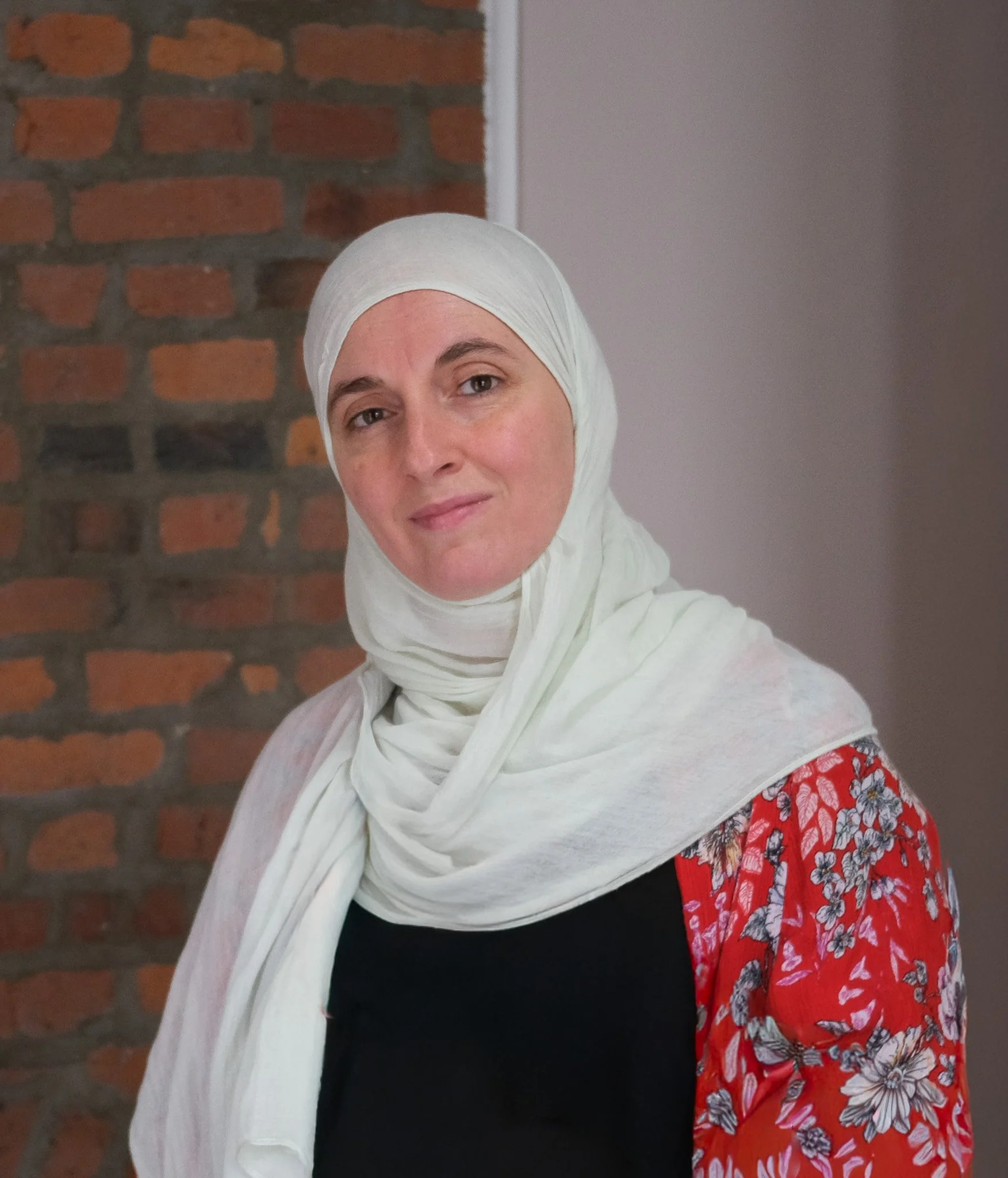 A woman wearing a white headscarf and a colorful red floral dress stands against a brick wall and gray background.