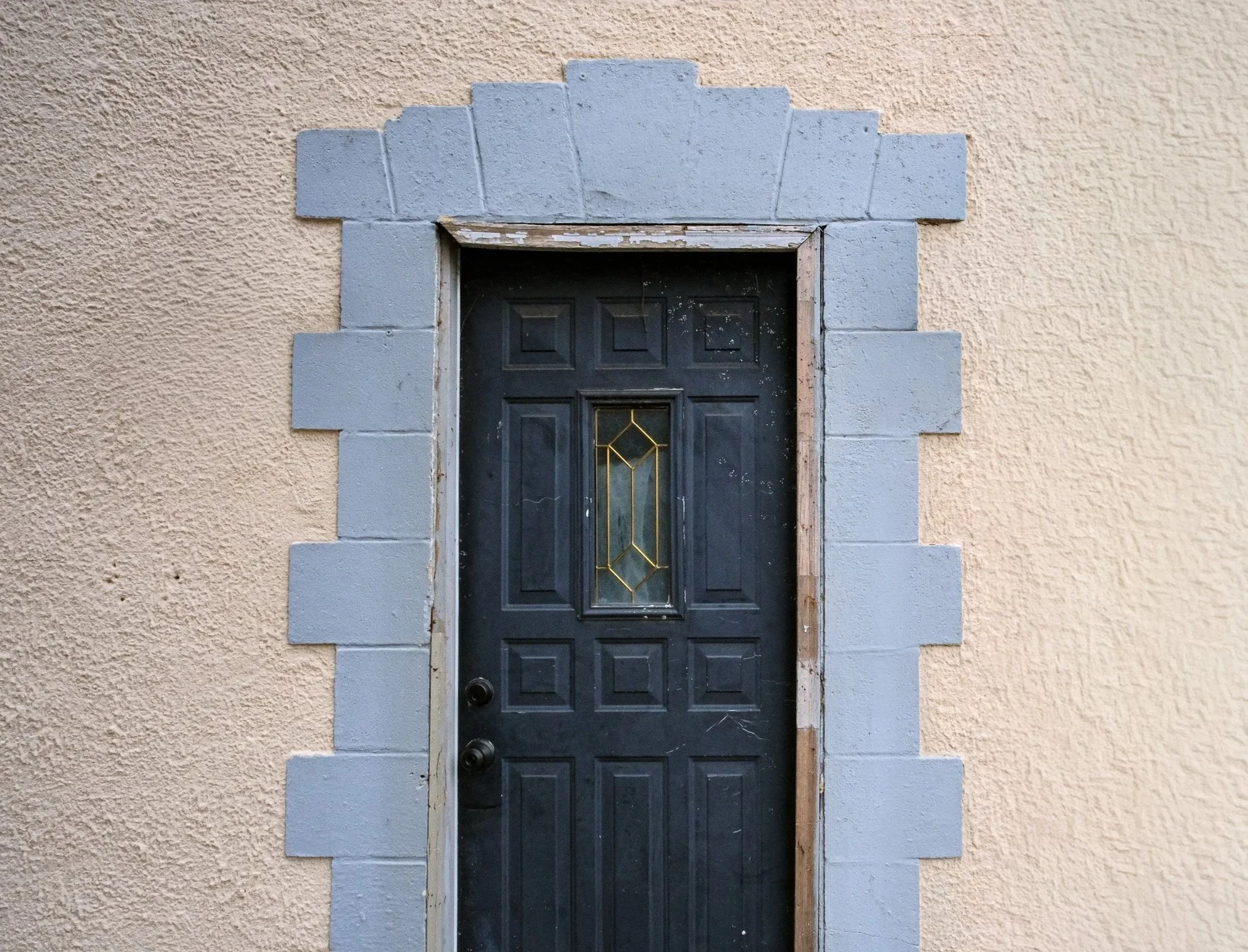 Front door with black color, decorative glass panel, surrounded by a beige stucco wall and a gray stone frame with an arch shape at the top.