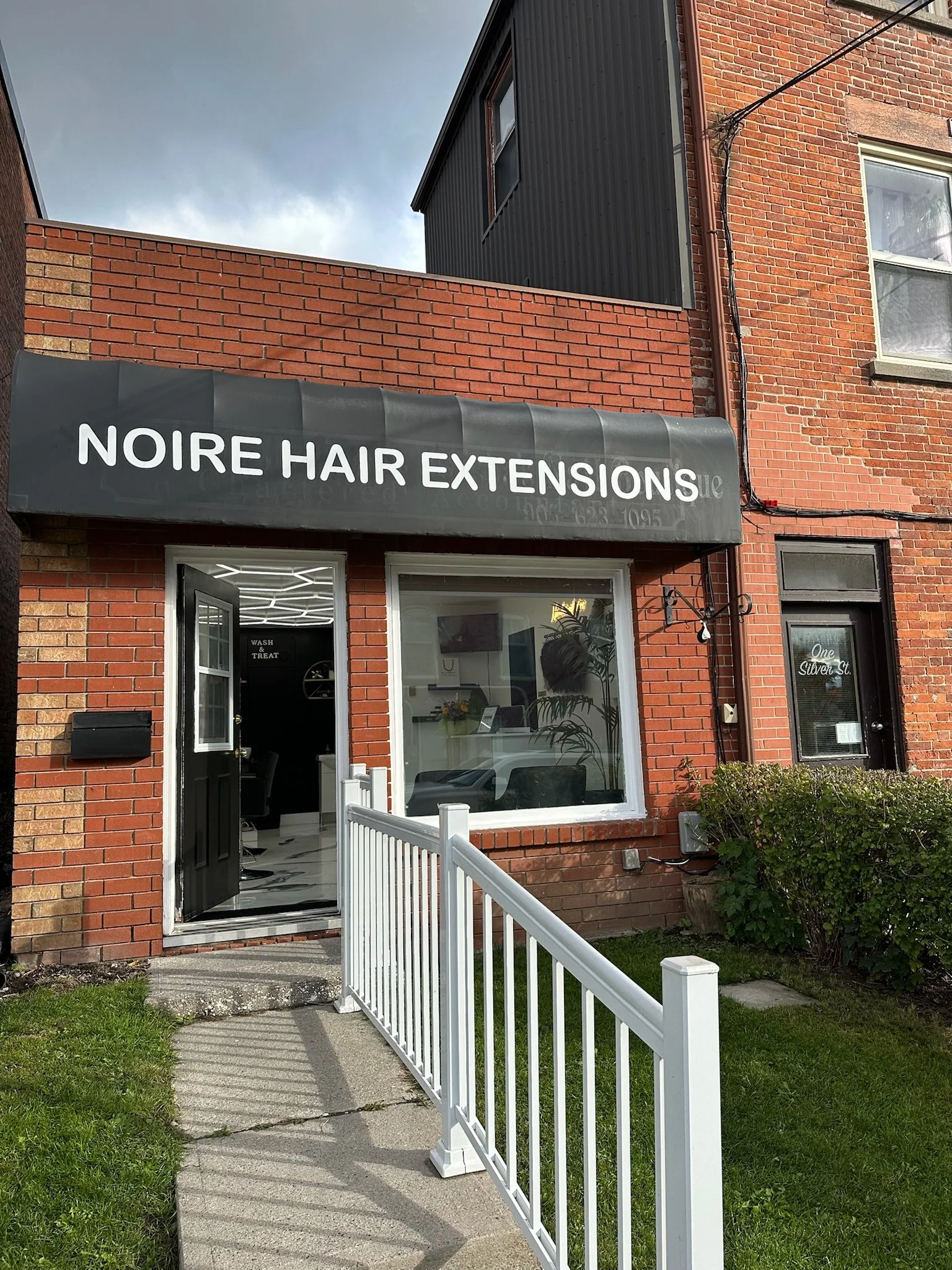Exterior view of a brick building with a black awning labeled 'Noire Hair Extensions' over the entrance. A white fence leads to the open door, revealing a modern interior.