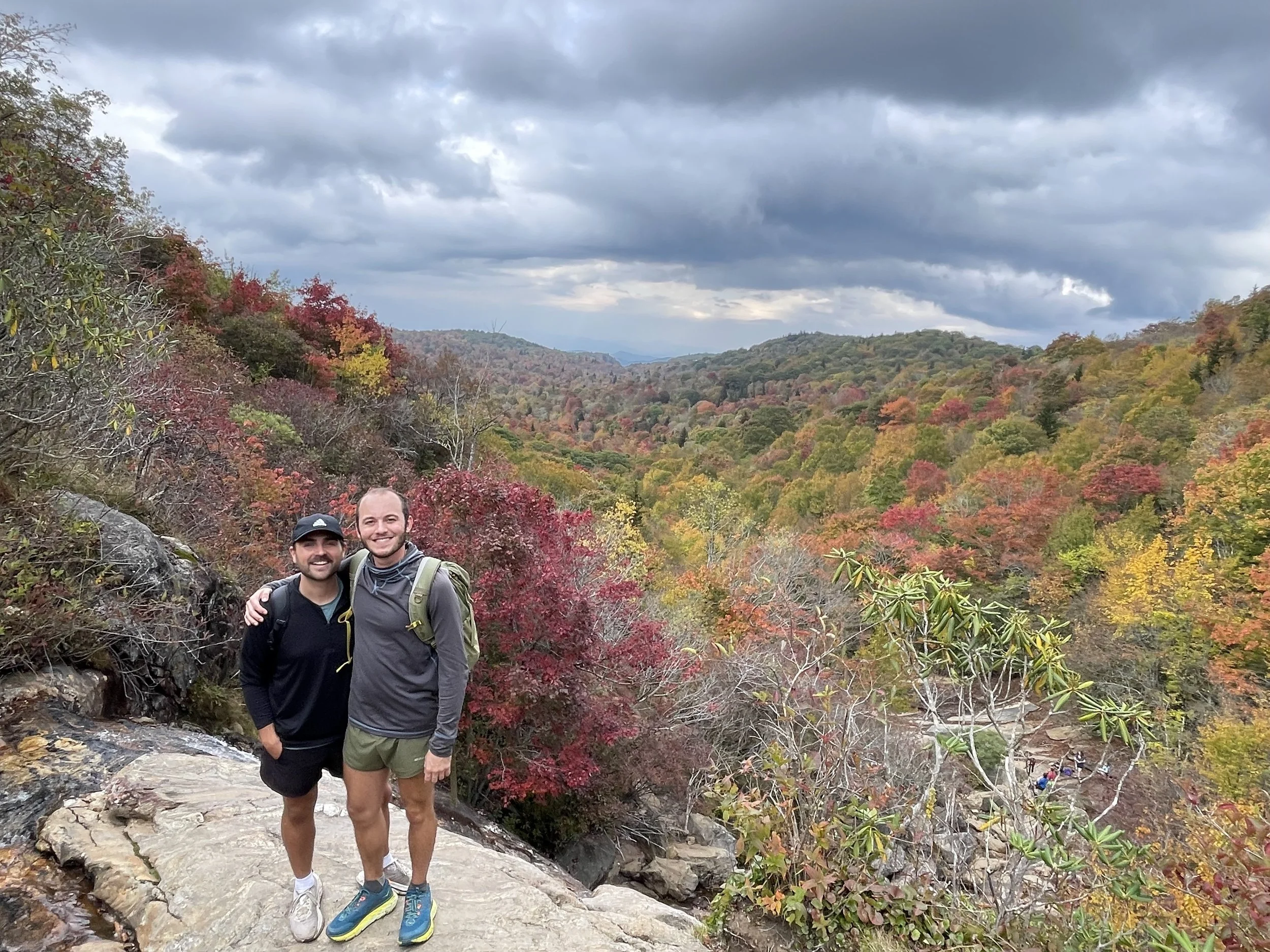 Two men smiling and posing with their arms around each other on a rocky trail in a mountain area during fall, with colorful autumn trees and a cloudy sky in the background.