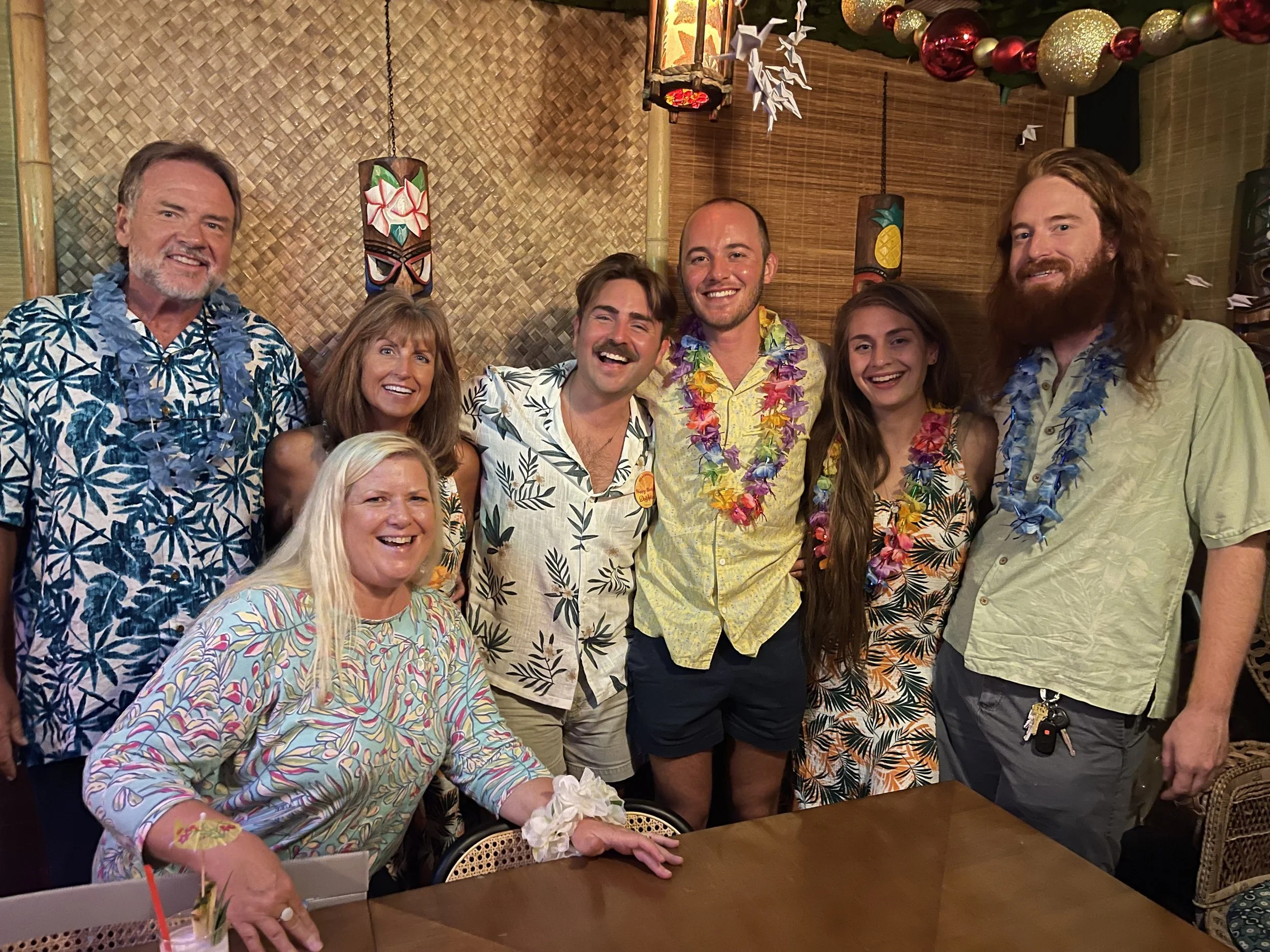 Group of people celebrating at a tropical-themed party, wearing colorful floral shirts and leis, with festive decorations in the background.