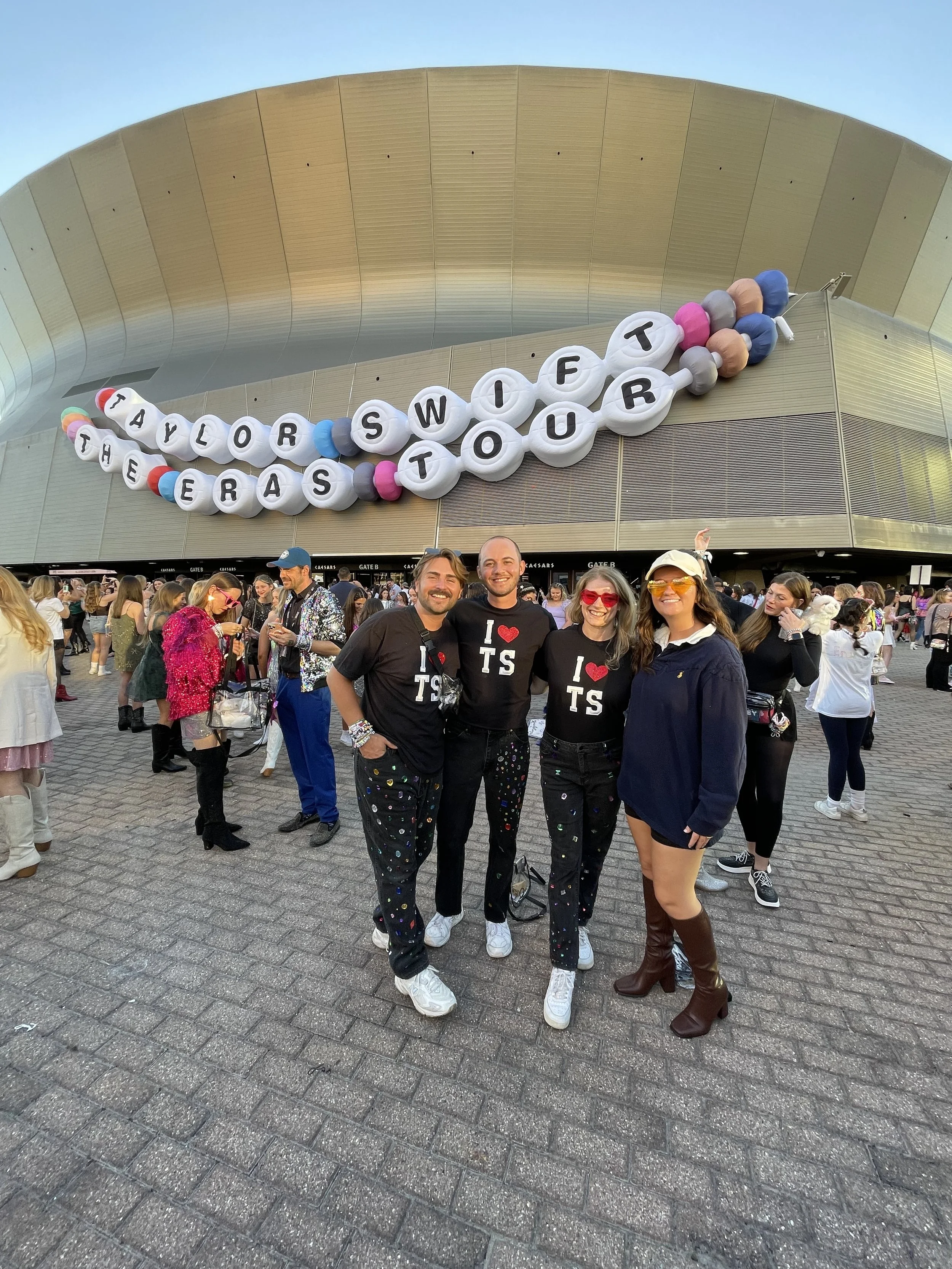 Group of four friends posing in front of a large balloon display that reads 'Taylor Swift The Eras Tour' outside of a stadium during daytime. The friends are smiling, dressed in casual and stylish outfits, with some wearing matching 'I ❤️ TS' shirts.