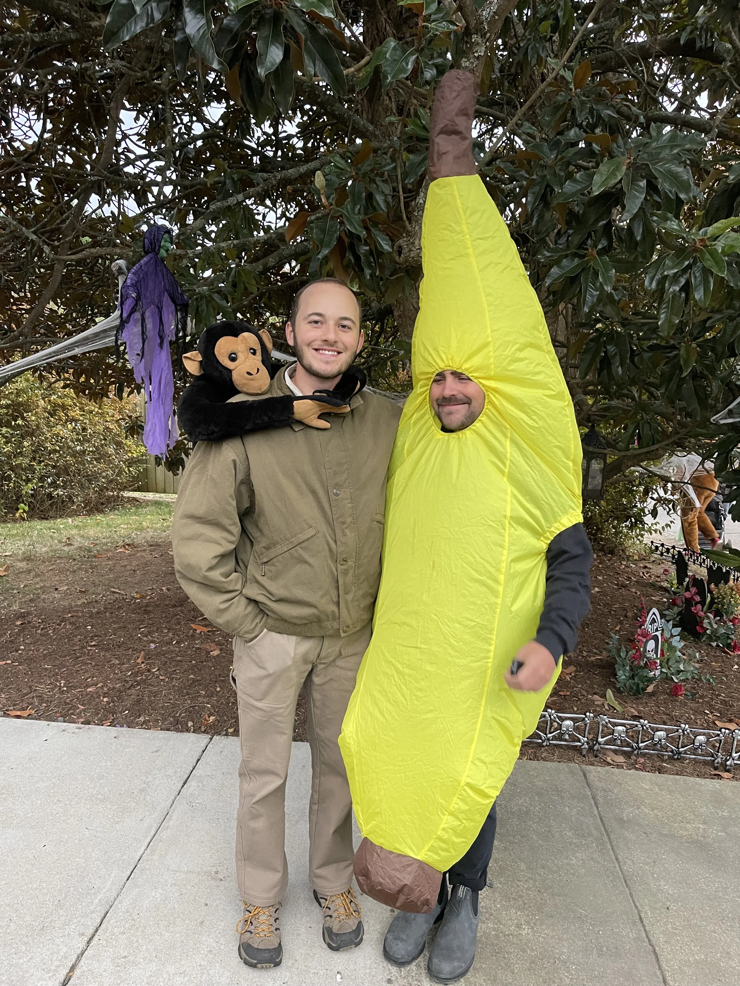 Two men in Halloween costumes standing outside on a sidewalk in front of a tree. One man is dressed as a banana, and the other is wearing a jacket with a monkey puppet on his shoulder. Halloween decorations, including purple spiders and flowers, are