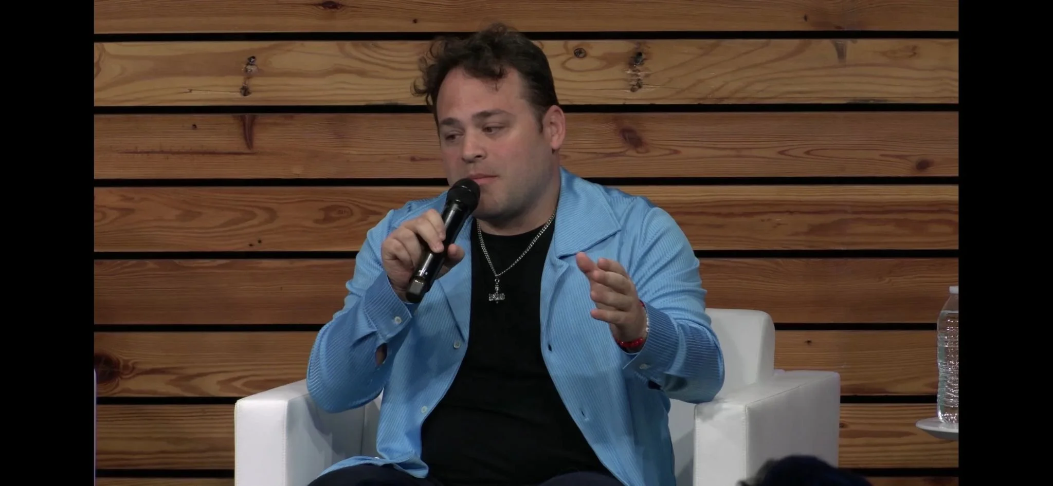A man in a blue jacket holding a microphone during a talk or interview, sitting in a white armchair with a wooden panel background.
