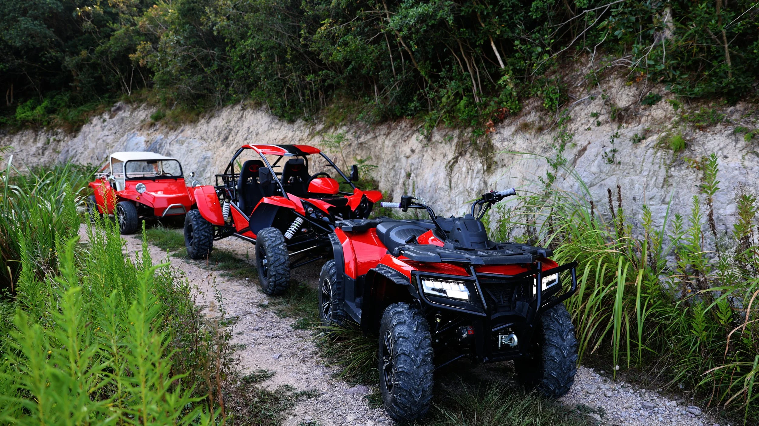 Three red off-road vehicles, including a quad bike, a dune buggy, and a go-kart, are parked on a dirt trail next to a rocky, shrub-lined hillside.