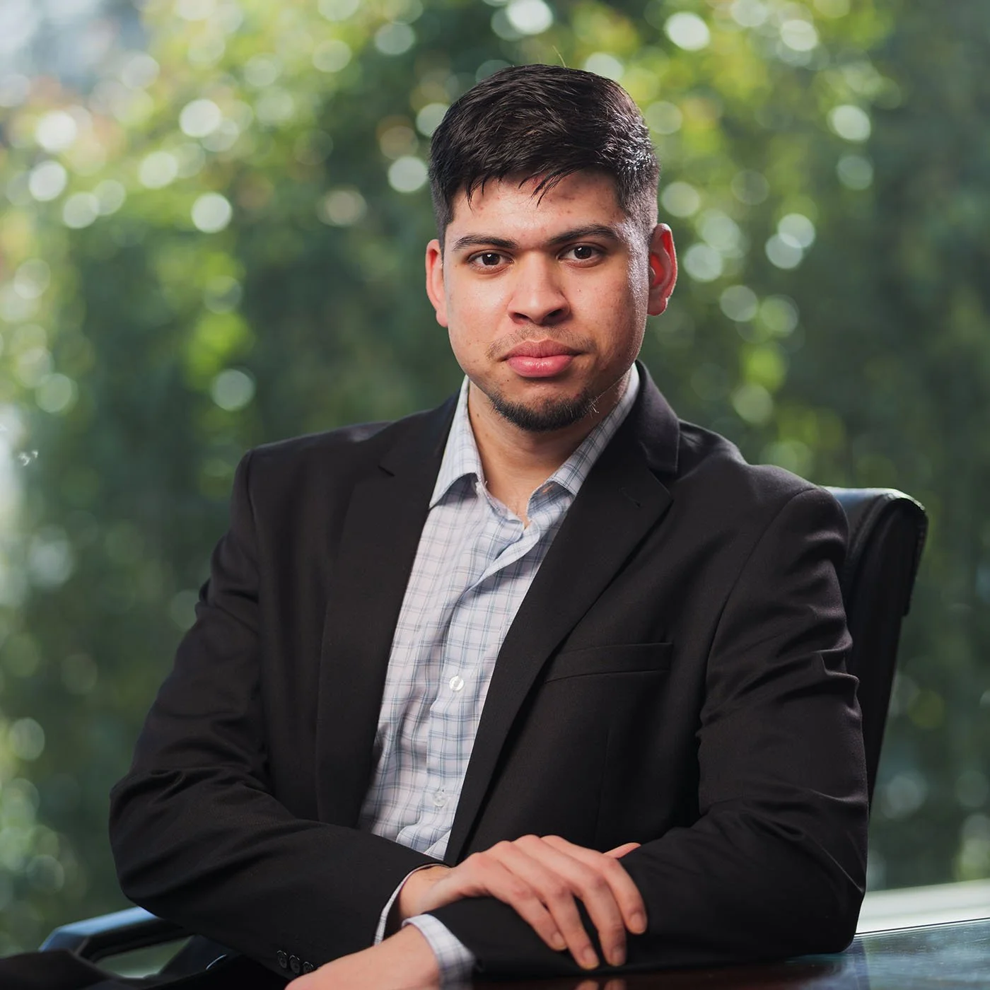 Marjuk Hossain with dark hair and a beard, wearing a black suit and a light checkered shirt, sitting in an office chair outdoors with a blurred green leafy background.