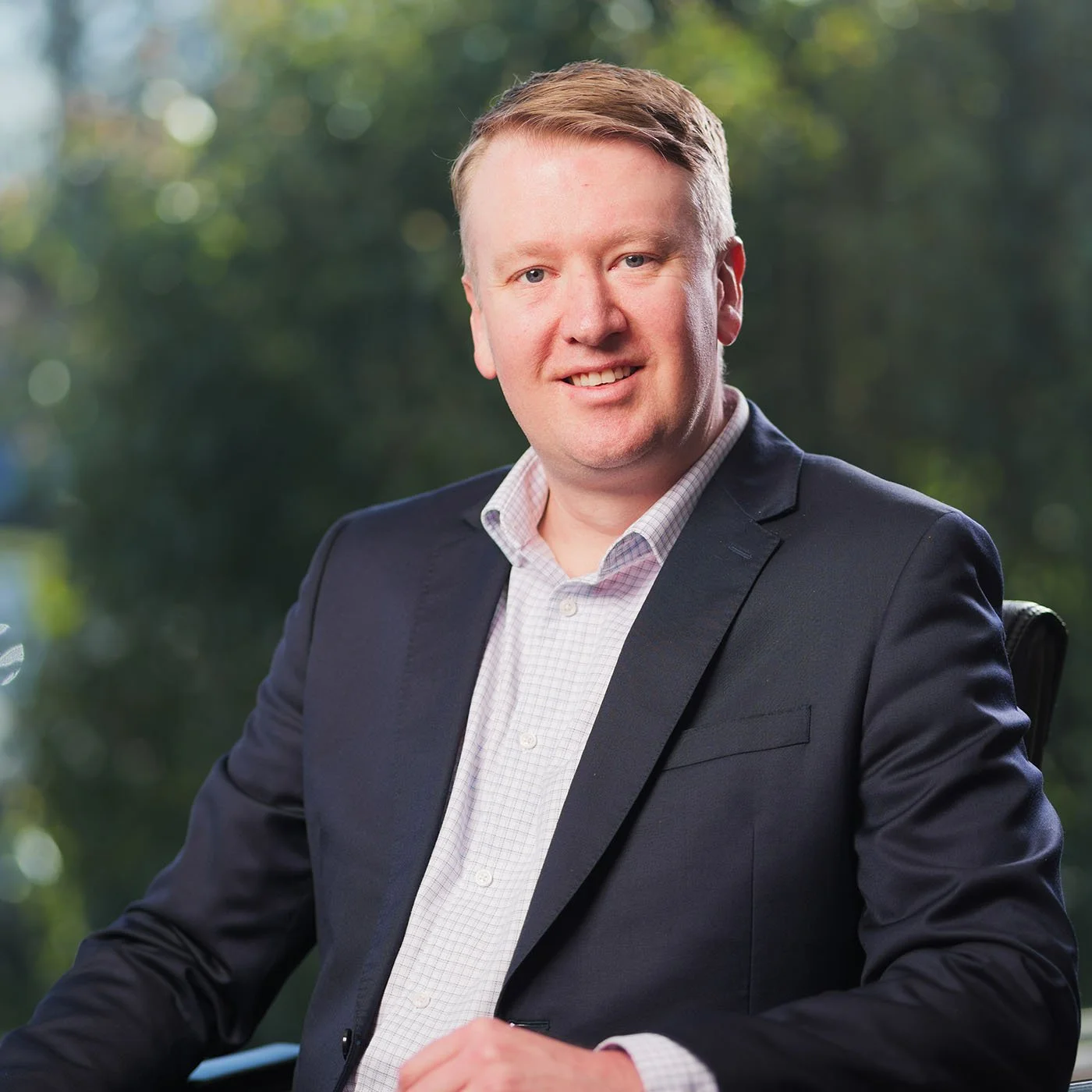 David Saynor - a smiling man with light skin, short light brown hair, wearing a dark suit jacket and white checkered shirt, sitting outdoors with a blurred green background.