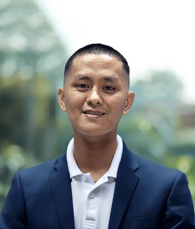 Headshot of a man with short dark hair, wearing a dark blue blazer and a white polo shirt, standing outdoors with blurred greenery in the background.