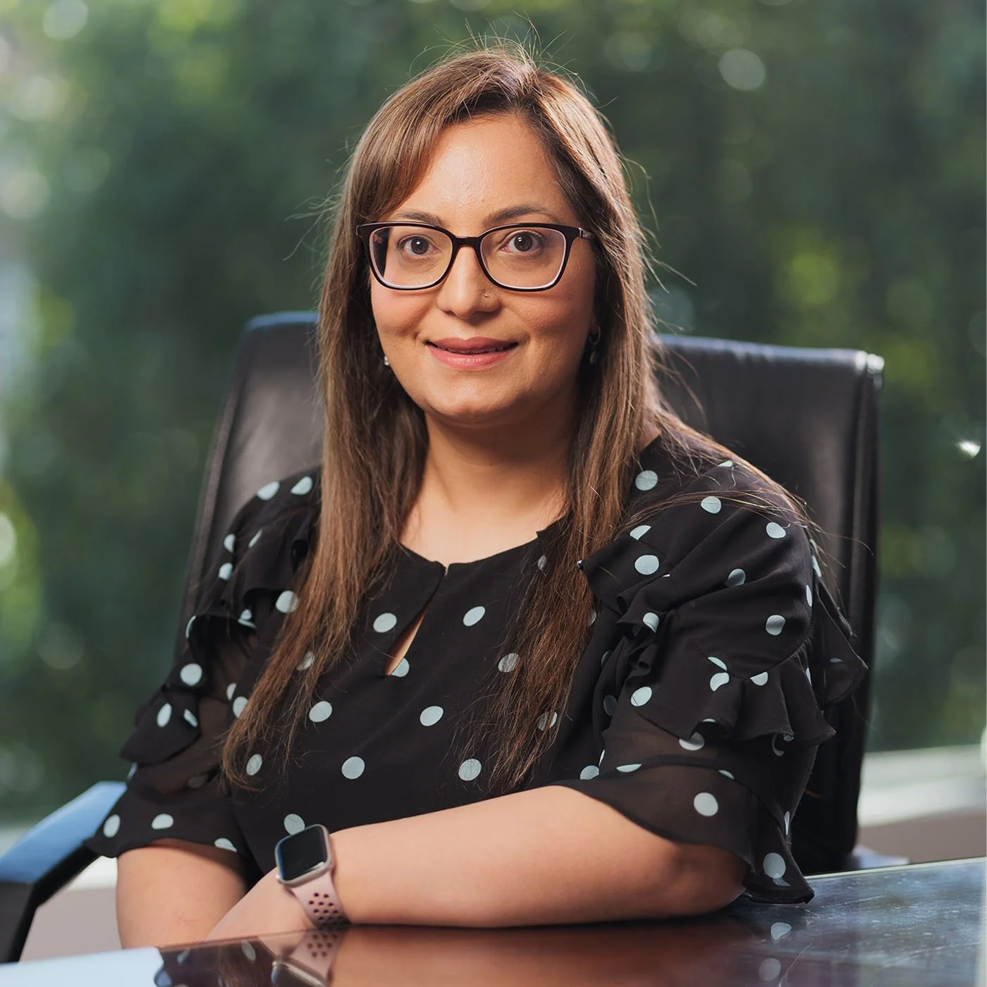 Ahla Qureshi with long brown hair and glasses, sitting at a desk with a black office chair, wearing a black dress with white polka dots and a smartwatch, outdoors with green blurred background.