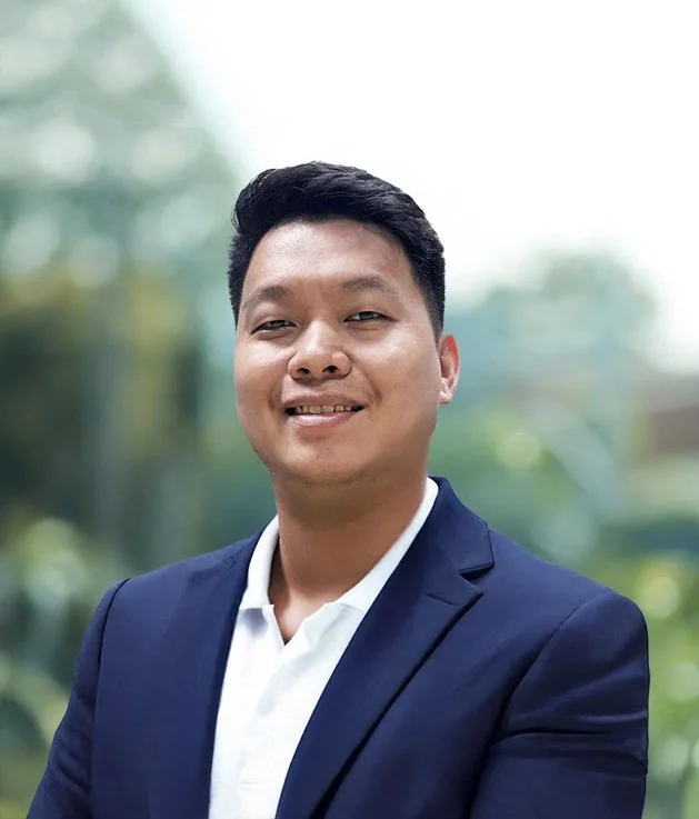 Portrait of a young man in a navy blazer and white shirt smiling outdoors with blurred green trees in the background.