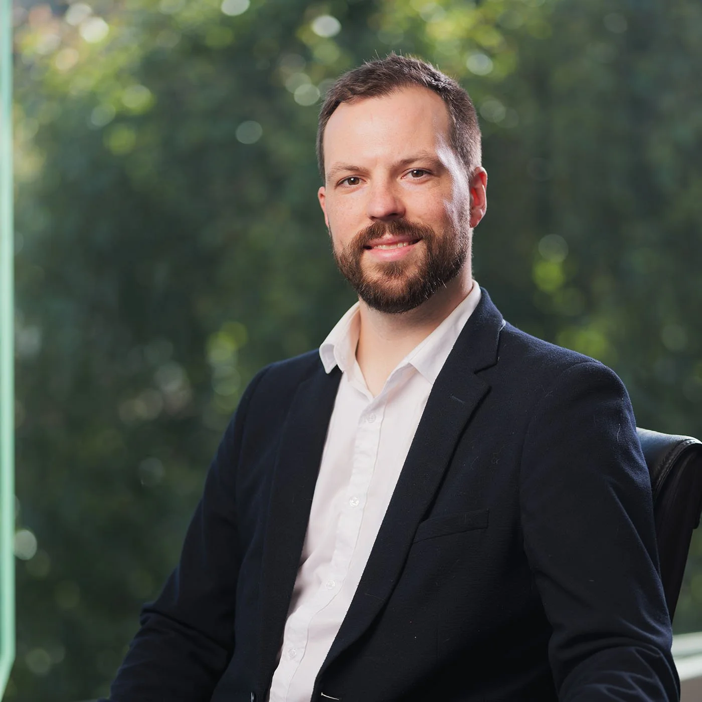 Matt Jacobs Financial Planner portrait - with a beard and short hair, wearing a white shirt and a dark blazer, seated outdoors with a blurred green background.