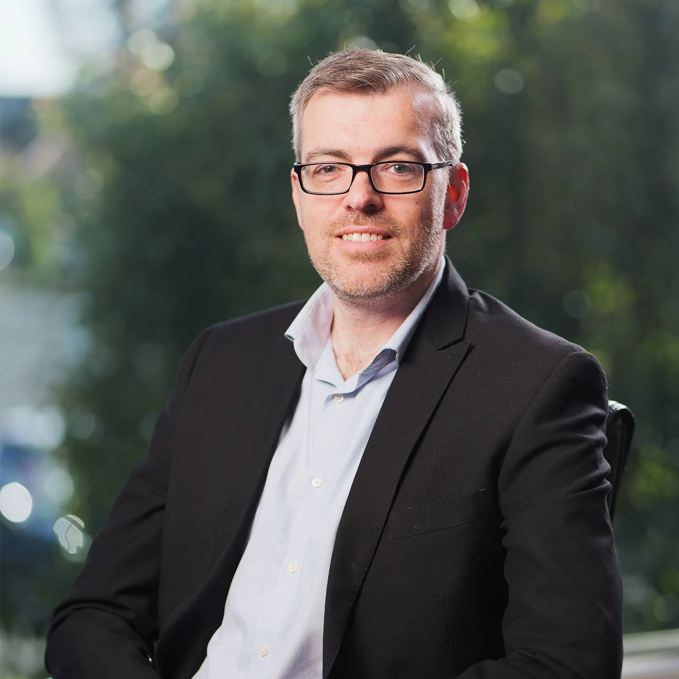 Brent Van Der Wel with glasses, short hair, and a beard, wearing a black blazer and a light blue shirt, sitting in front of a blurred background of green trees and sunlight.