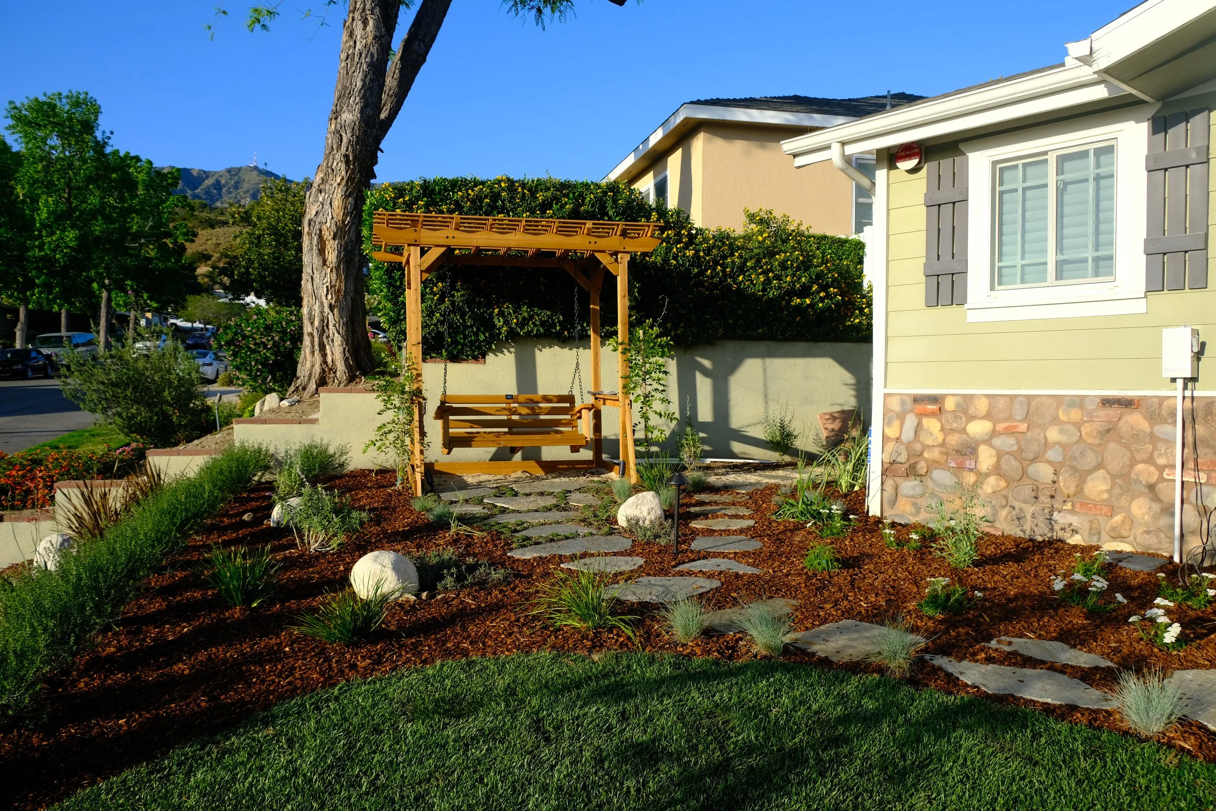 A landscaped front yard with a stone pathway leading to a wooden bench swing under a pergola, surrounded by various plants and mulch, with a house and trees in the background.