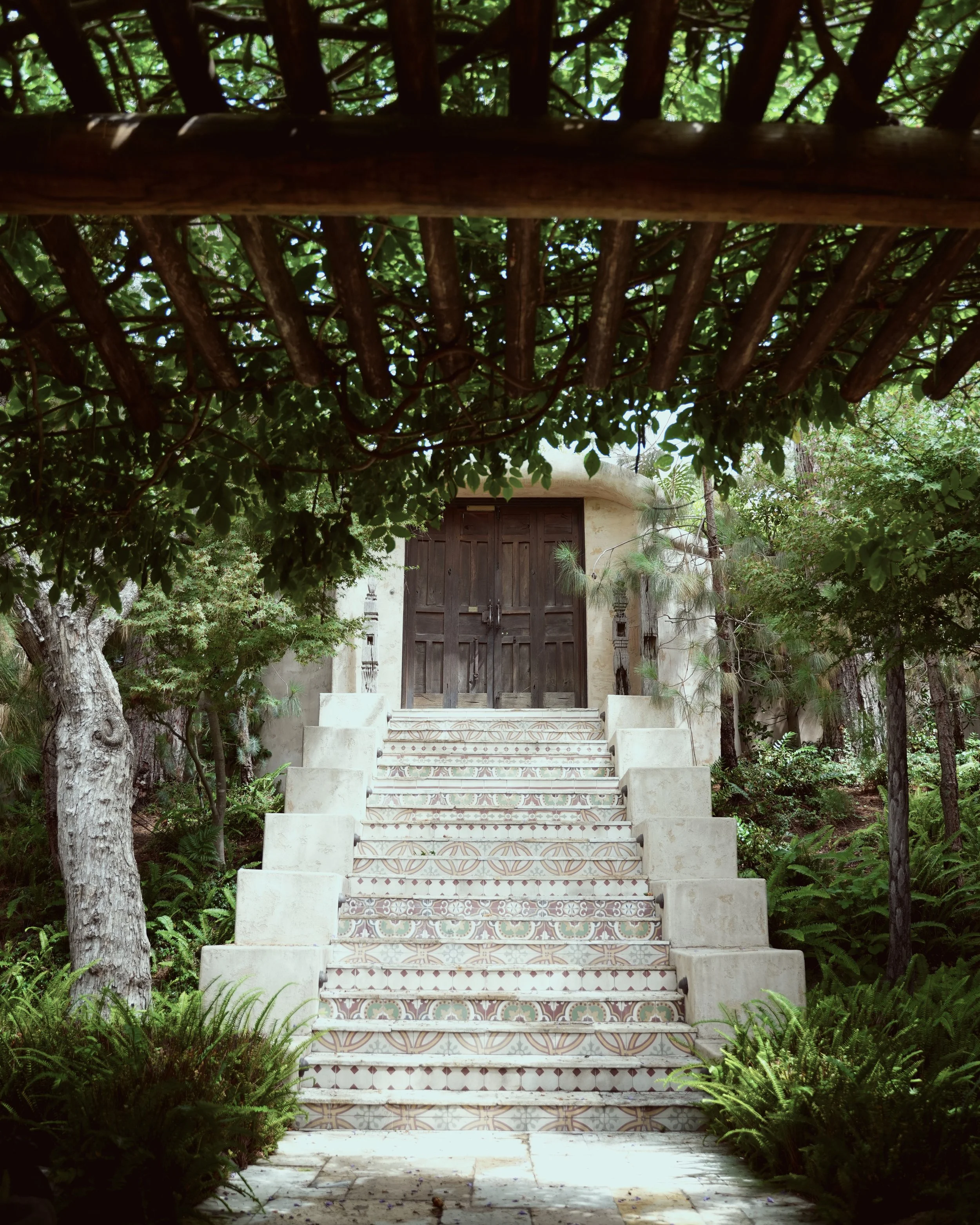 A set of decorative stairs leading up to a wooden door, surrounded by lush green trees and vegetation, in a garden setting.
