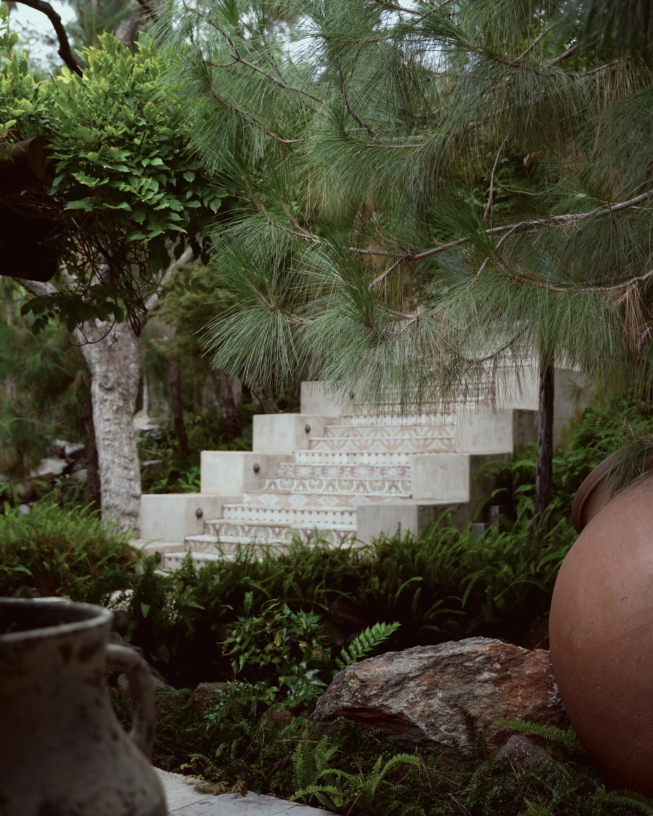 A stone staircase with decorative tiles, surrounded by lush green foliage, trees, and large pots in a garden setting.