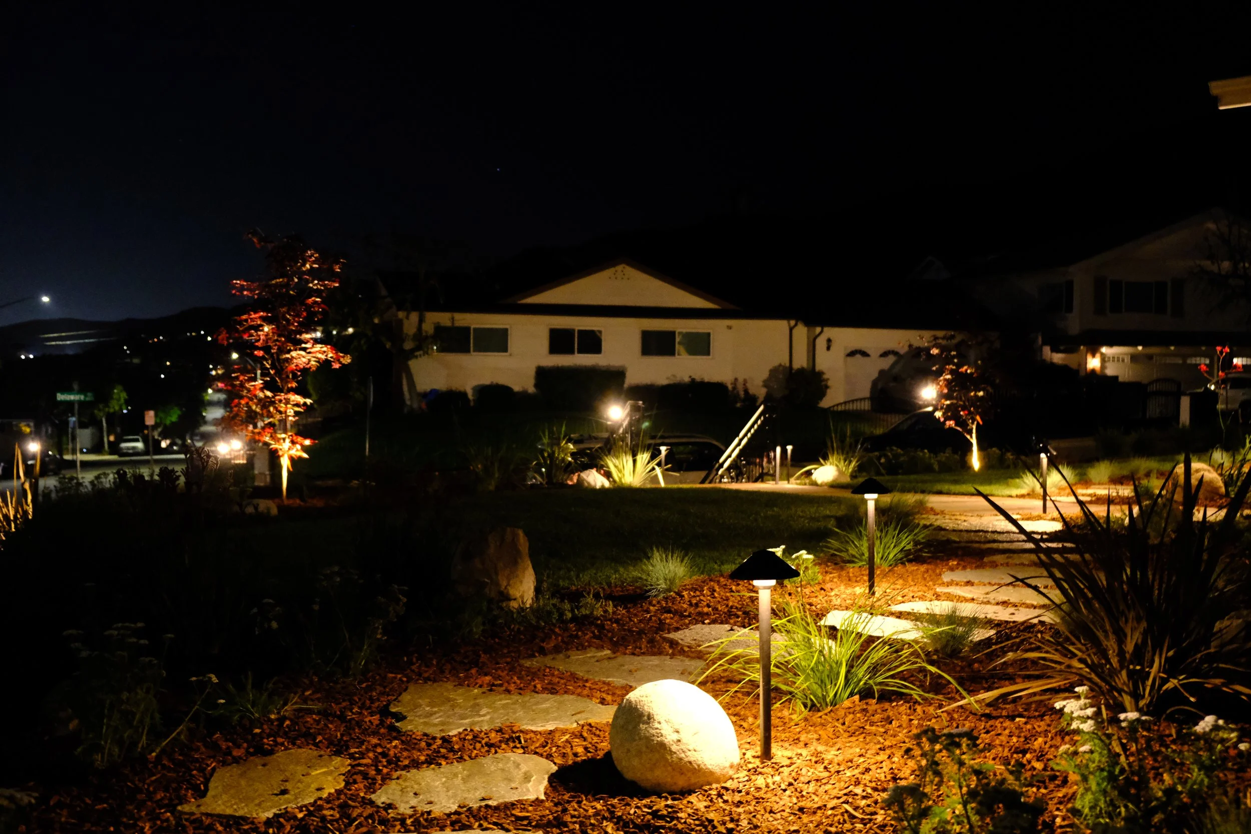 Nighttime view of a landscaped front yard with illuminated pathway, rocks, small trees, bushes, and houses in the background.