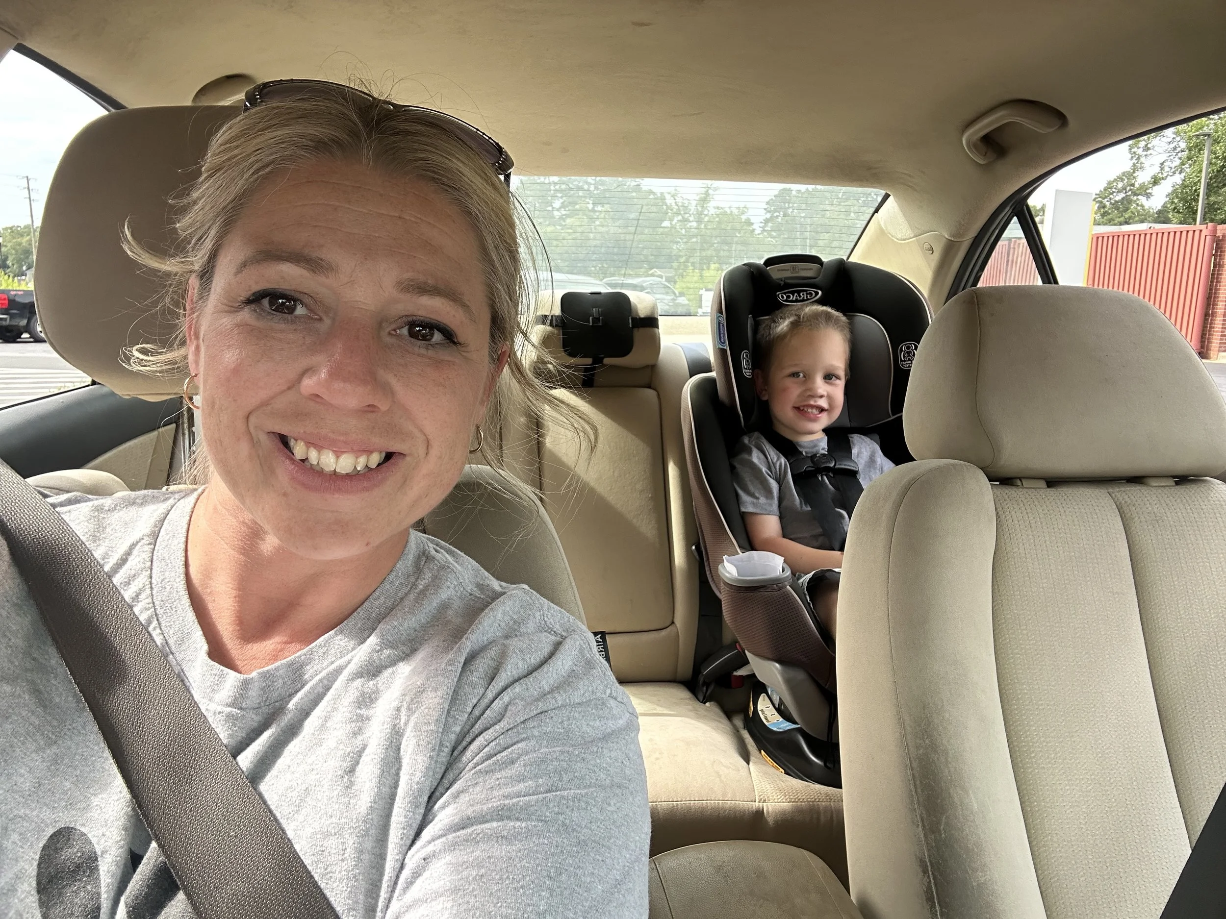 A woman taking a selfie inside a car with a young boy sitting in a car seat in the back. The woman is smiling, wearing a gray shirt and has sunglasses on her head. The boy is smiling, wearing a gray shirt, and secured in a child safety seat.