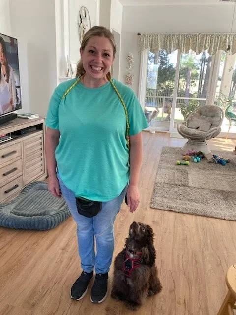 A woman with a yellow measuring tape around her neck smiling at the camera, standing in a living room with her brown dog sitting beside her.