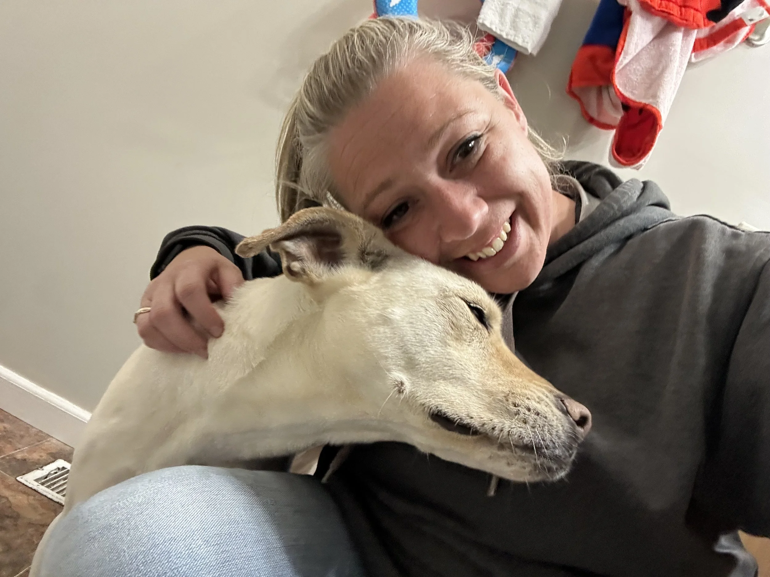A woman smiling and hugging a dog with closed eyes inside a room, with laundry hanging in the background.