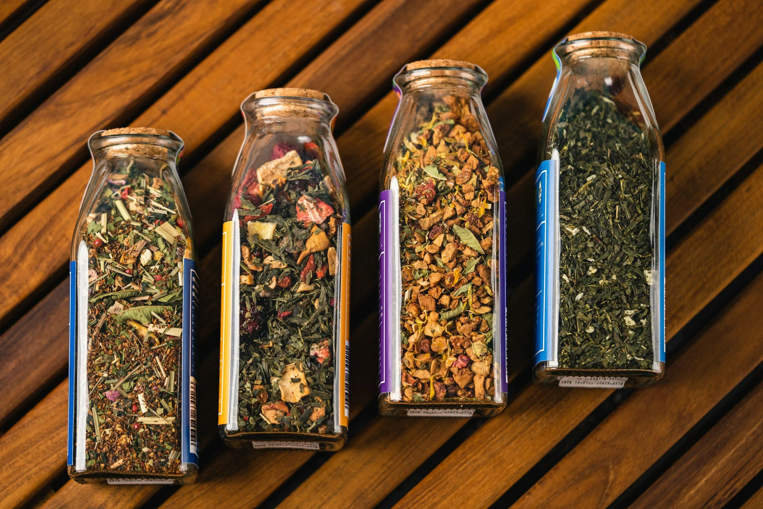 Four glass spice jars with various dried herbs and spices on a wooden surface.