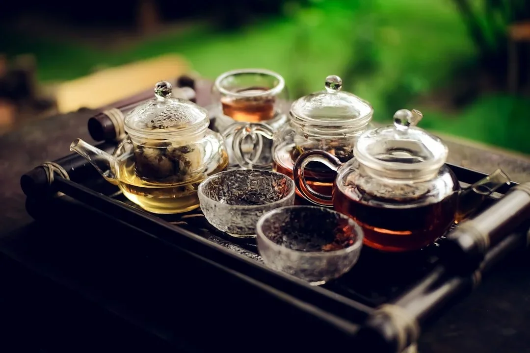 Set of glass teapots with different types of tea and two small glass cups on a tray, outdoor garden background.