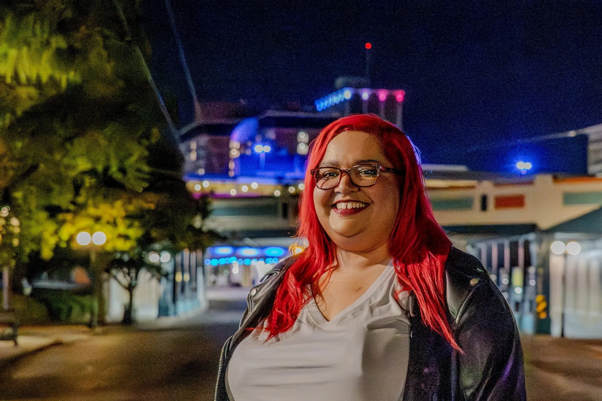 A person with red hair, glasses, and a black leather jacket standing outdoors at night, smiling, with a brightly lit city scape in the background.