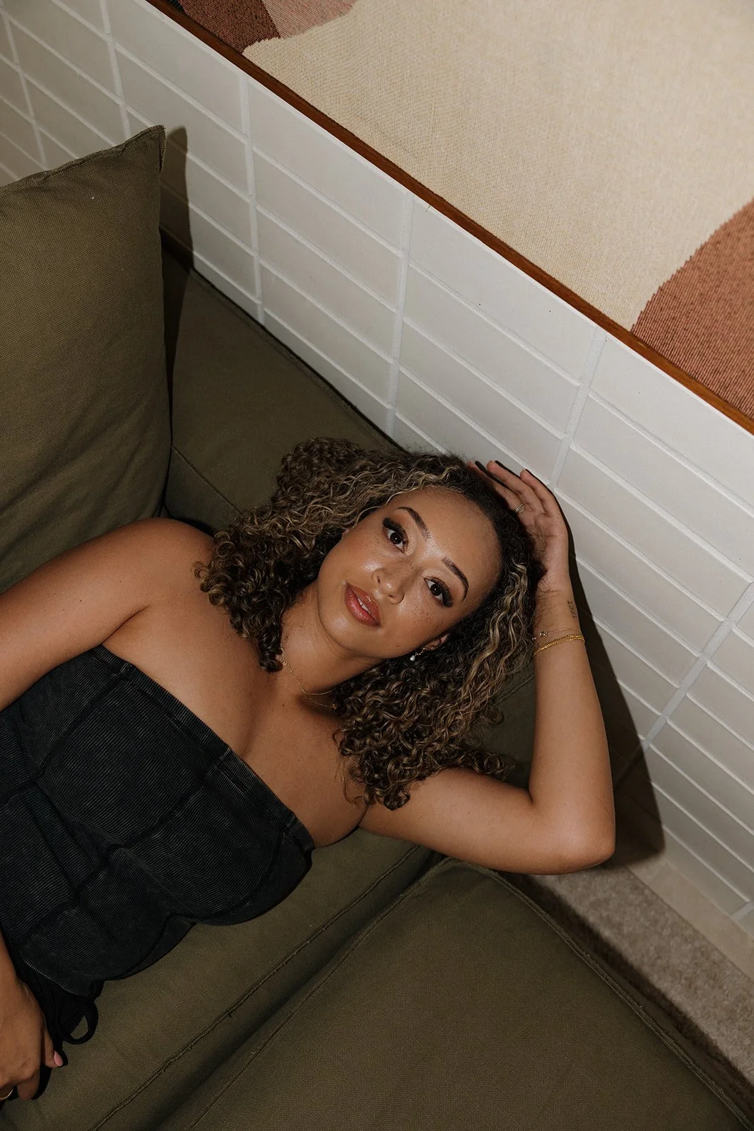 A woman with curly hair lying on a green sofa, resting her head on her hand, looking at the camera.