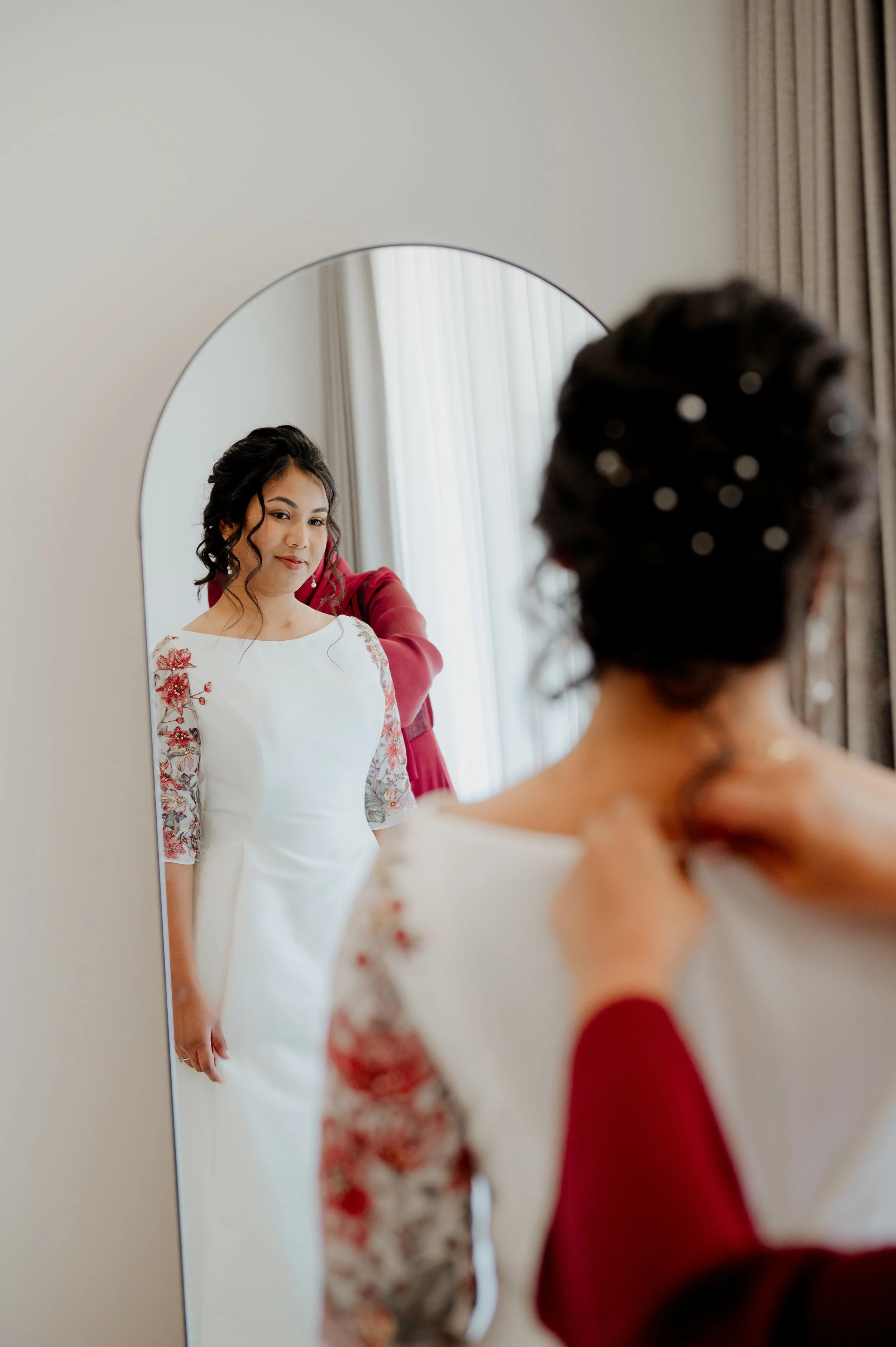 A woman wearing a white dress with floral embroidery on the sleeves looks at her reflection in a mirror as another woman helps her with her outfit, possibly pre-wedding or special occasion clothes.
