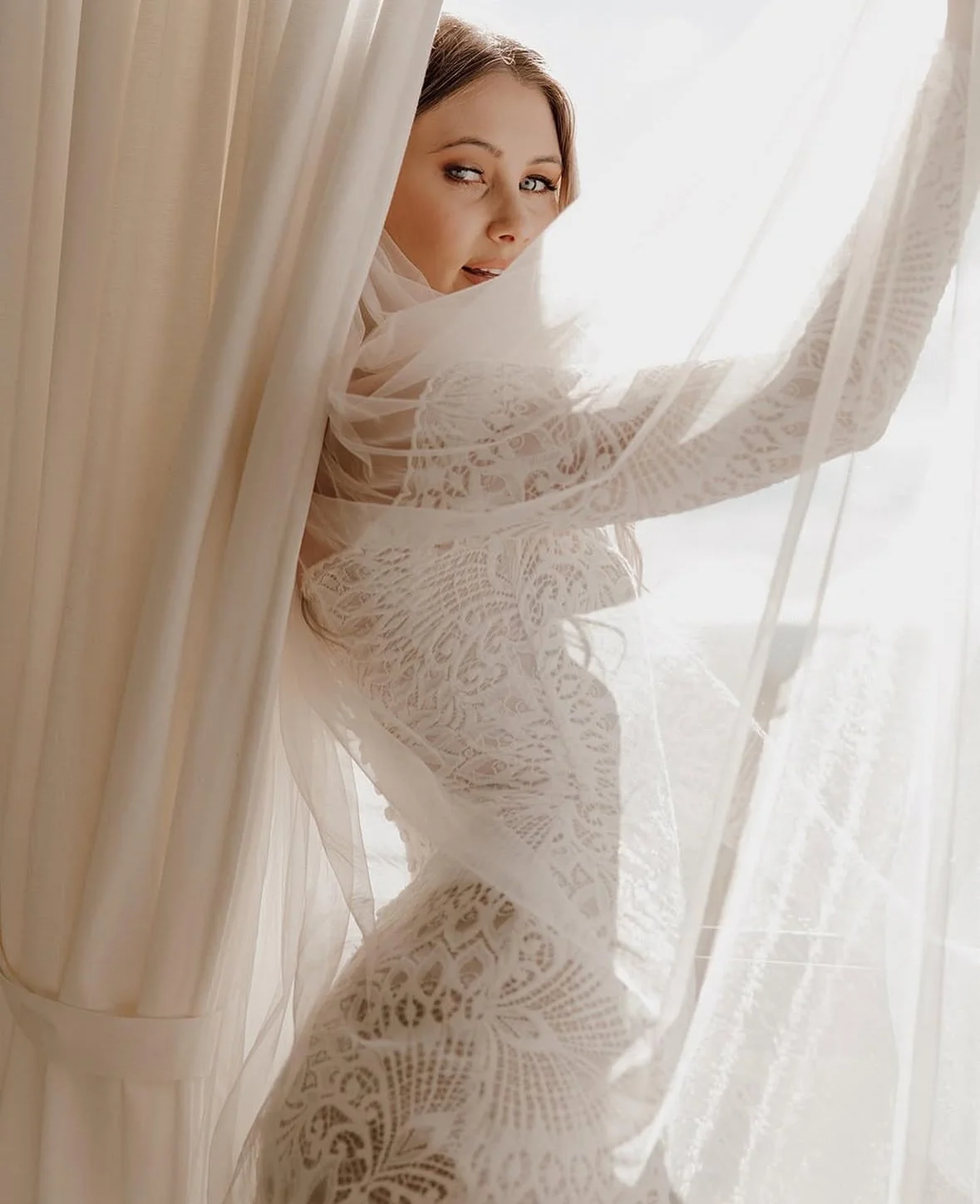 A woman with light brown hair is peeking around a sheer, lace curtain. Sunlight streams in through the window behind her, creating a soft, bright atmosphere.