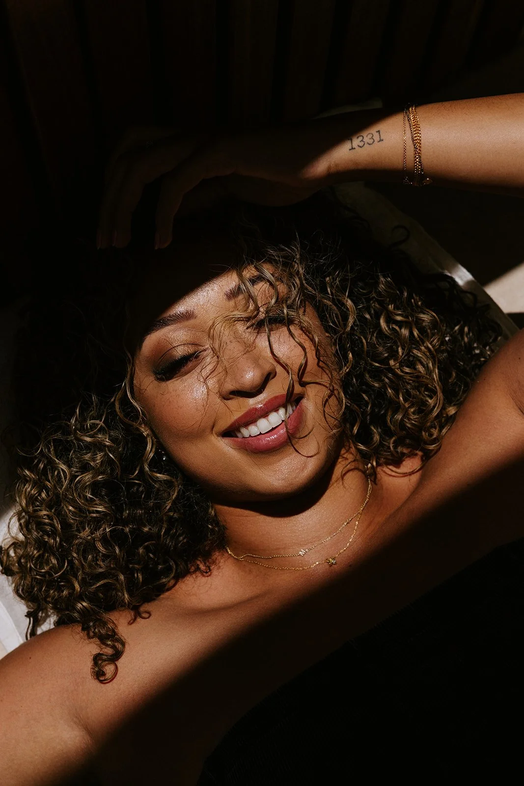 Close-up of a woman with curly hair smiling with her eyes partly closed, illuminated by sunlight, wearing jewelry including layered necklaces and bracelets.