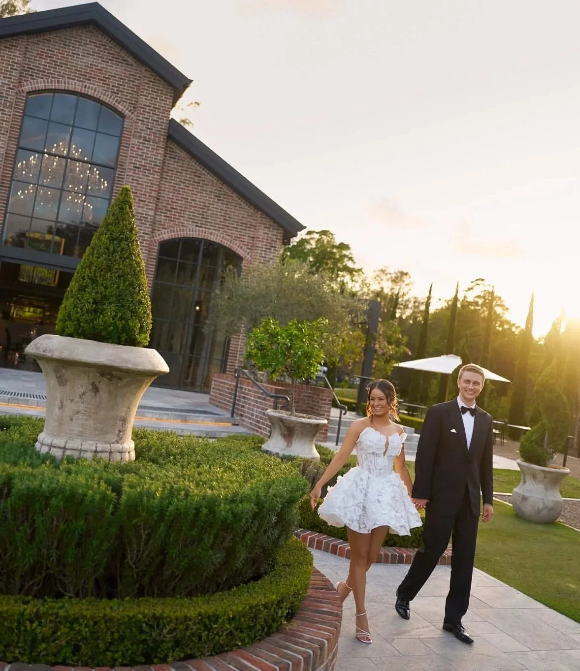 A young woman and man dressed in formal attire walk hand in hand outside a brick building with large windows, greenery, and large decorative planters during sunset.