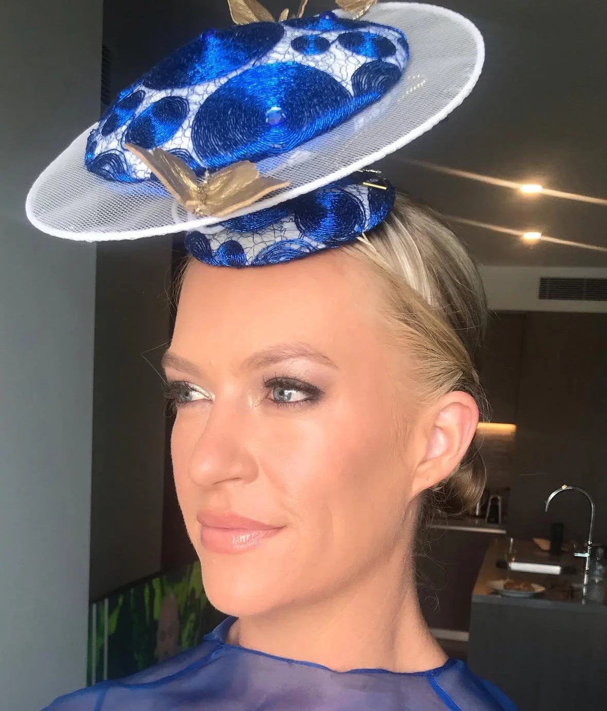Woman wearing a stylish fascinator hat with blue and white mesh, lace, and gold accents, indoors with a modern kitchen background.