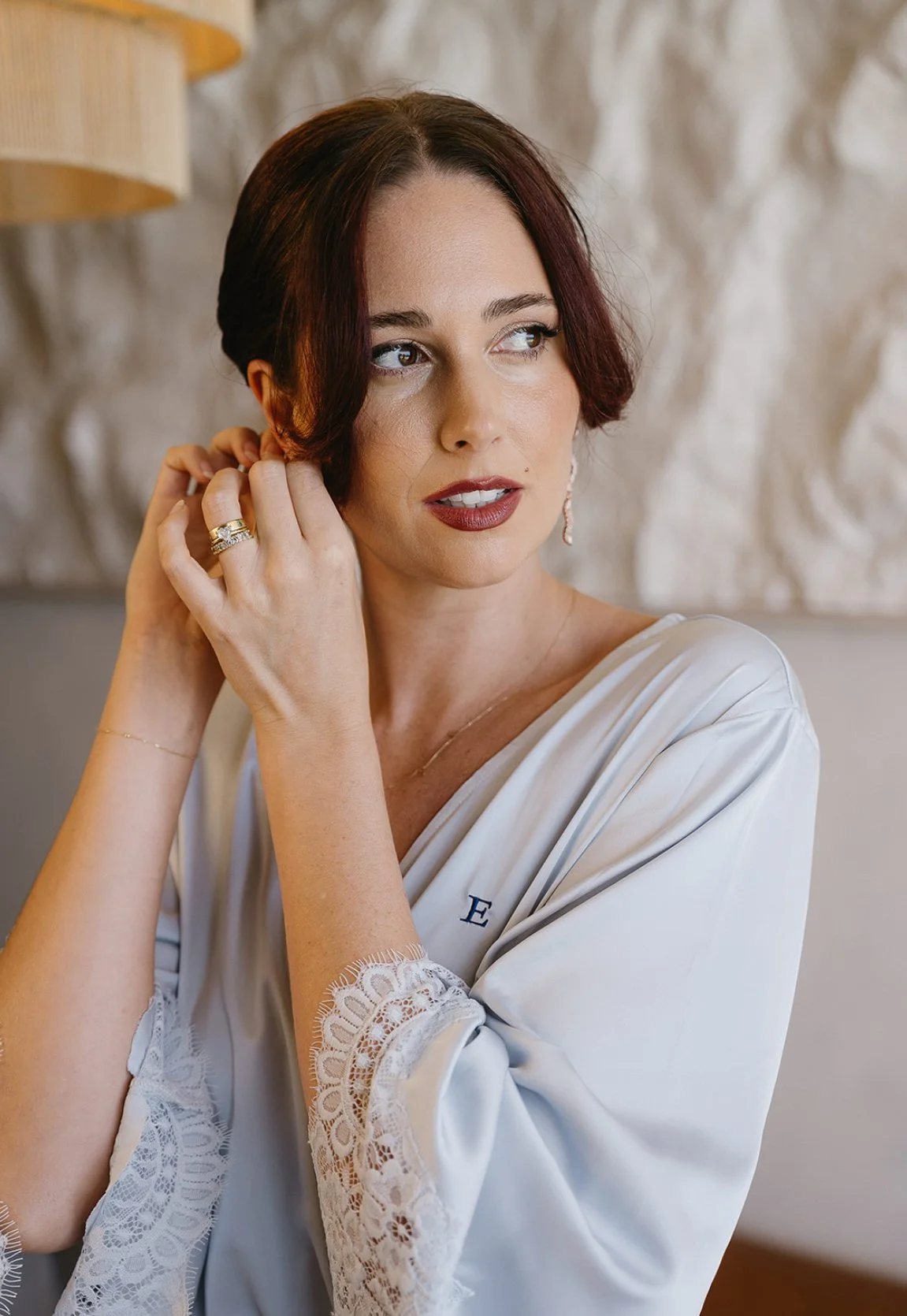 A woman with short dark hair, wearing makeup and jewelry, is putting on an earring while dressed in a white satin robe with lace cuffs, in a softly lit room.