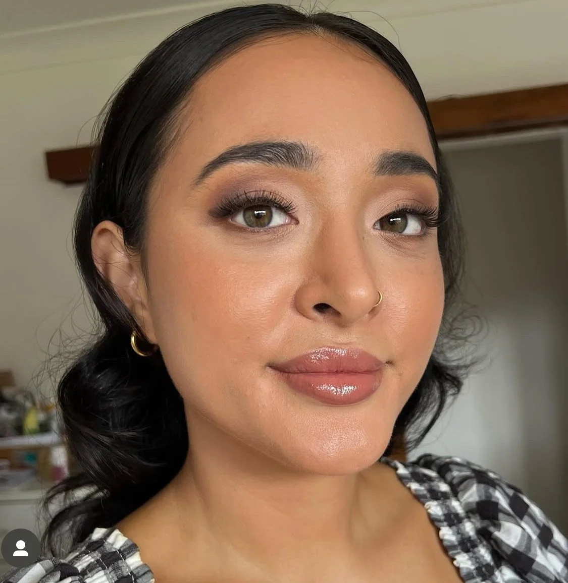A woman with long dark hair, wearing makeup with defined eyebrows, eye shadow, mascara, glossy lips, a small hoop earring, a nose ring, and a checkered top. Interior background with a wooden beam and blurred objects.