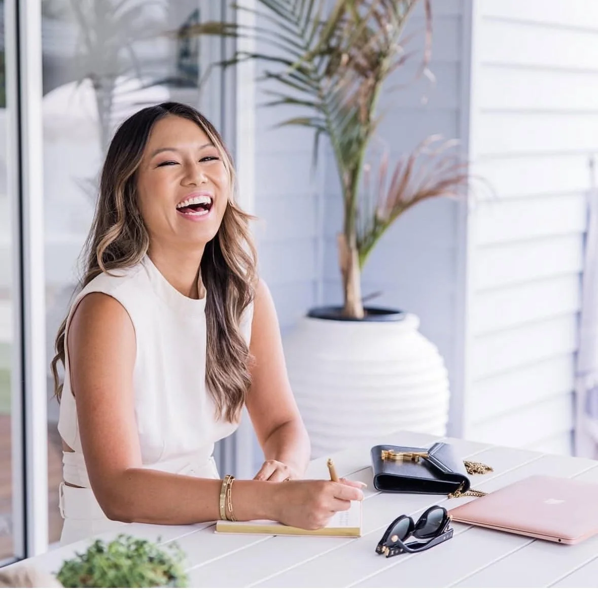 A woman smiling and laughing while sitting at a table with a notebook, sunglasses, a black purse, and a closed pink laptop.