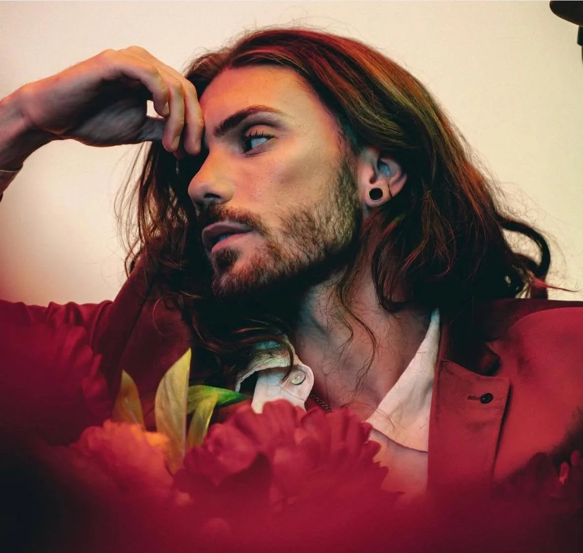 A man with long, dark hair and beard, wearing a red jacket and white shirt, posing with his hand on his forehead, surrounded by red flowers.