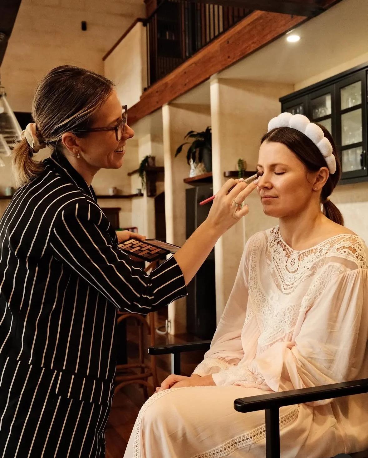 A woman in a black and white striped blazer is applying makeup to a seated woman with dark hair and a white lace headband, sitting in a beige dress with lace details, in a cozy home setting.