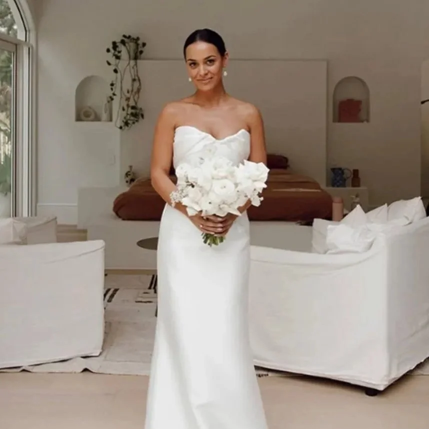 A woman in a white strapless wedding dress holding a bouquet of white flowers in a bright, modern room.