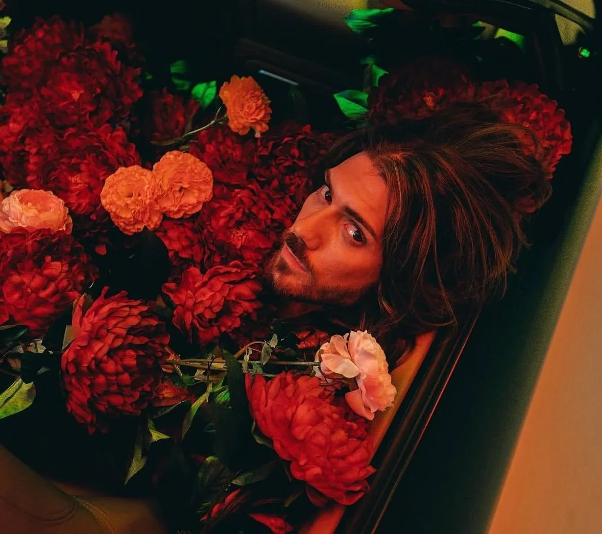 A man with long hair and a beard lying down among red and orange flowers, looking at the camera.